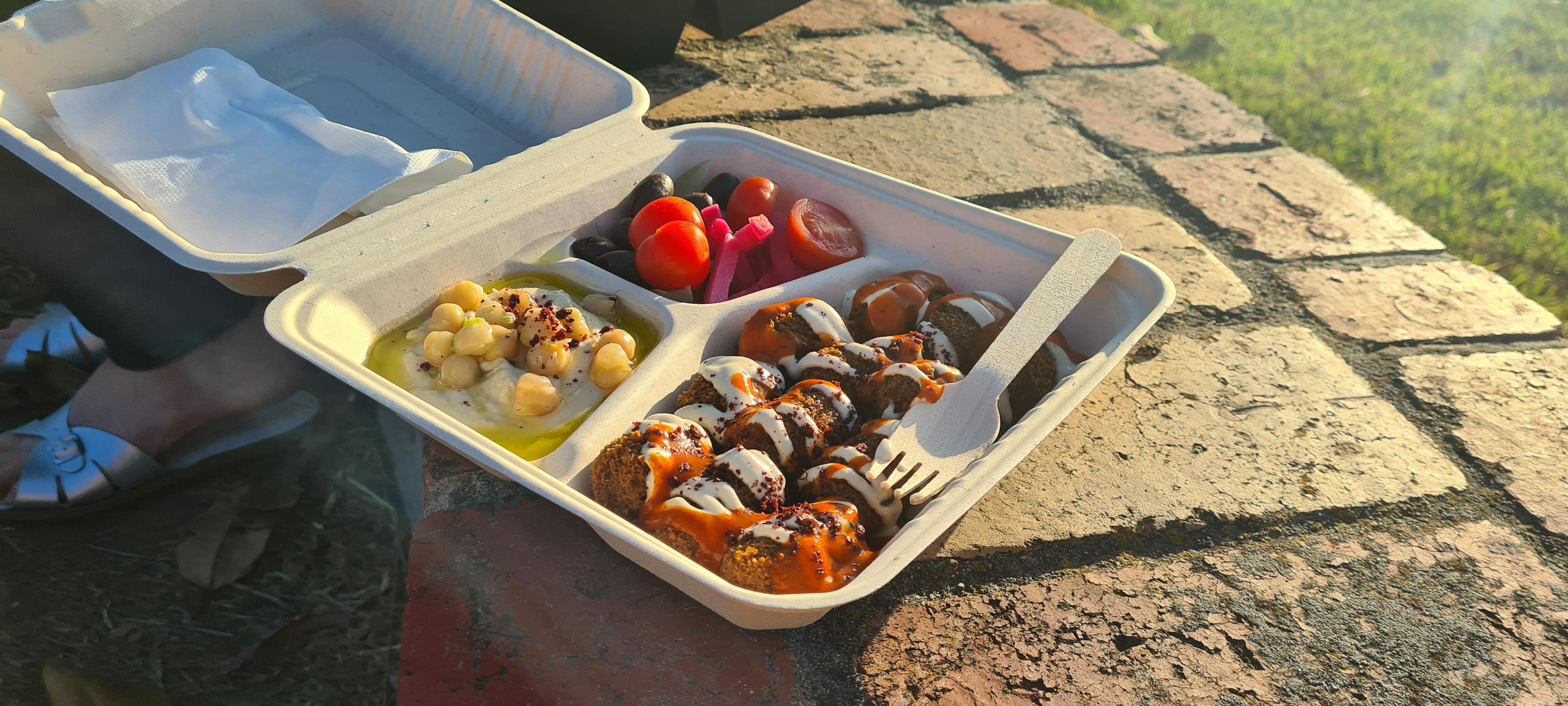 Many falafel, coated in garlic sauce and chilli sauce, next to hummus with whole chickpeas and pickles.