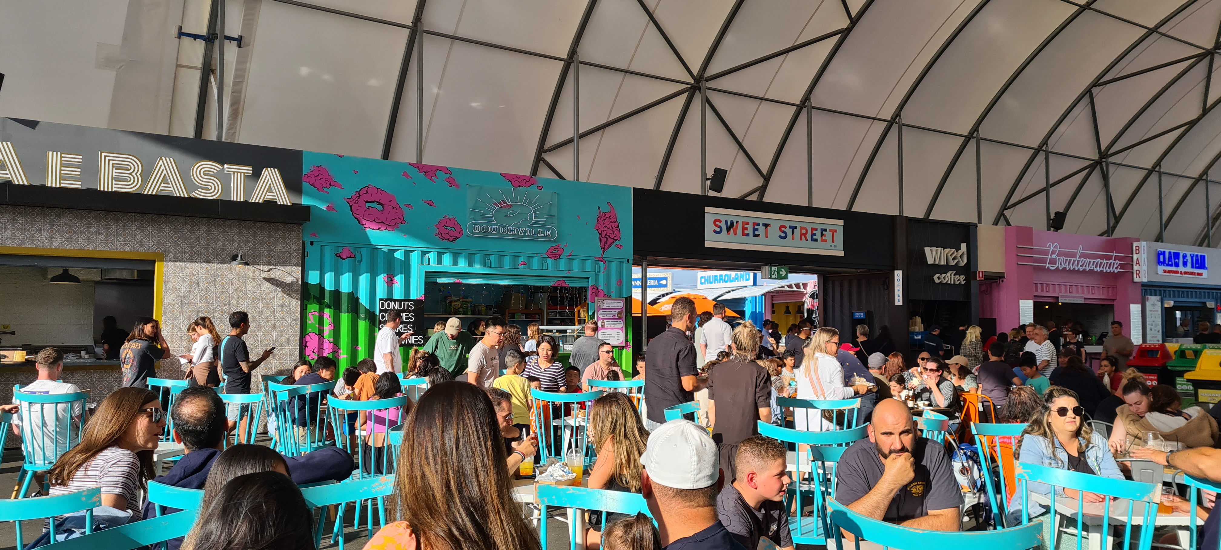 Multiple shipping container side-by-side, painted and decorated, selling different foods, with people sitting and eating at tables in a central area.