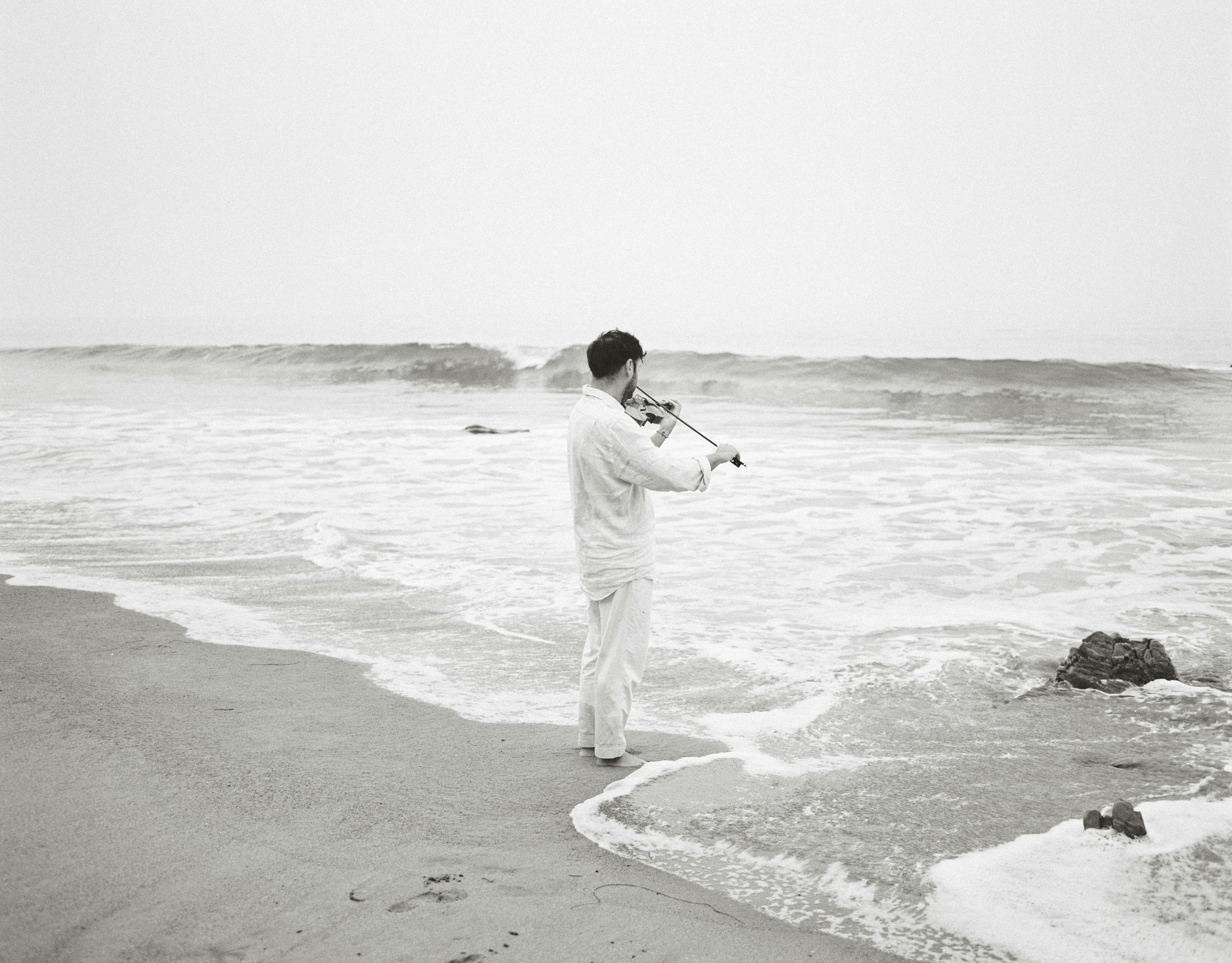 Bryan Senti playing violin in front of the Pacific Ocean. Black and white photo.