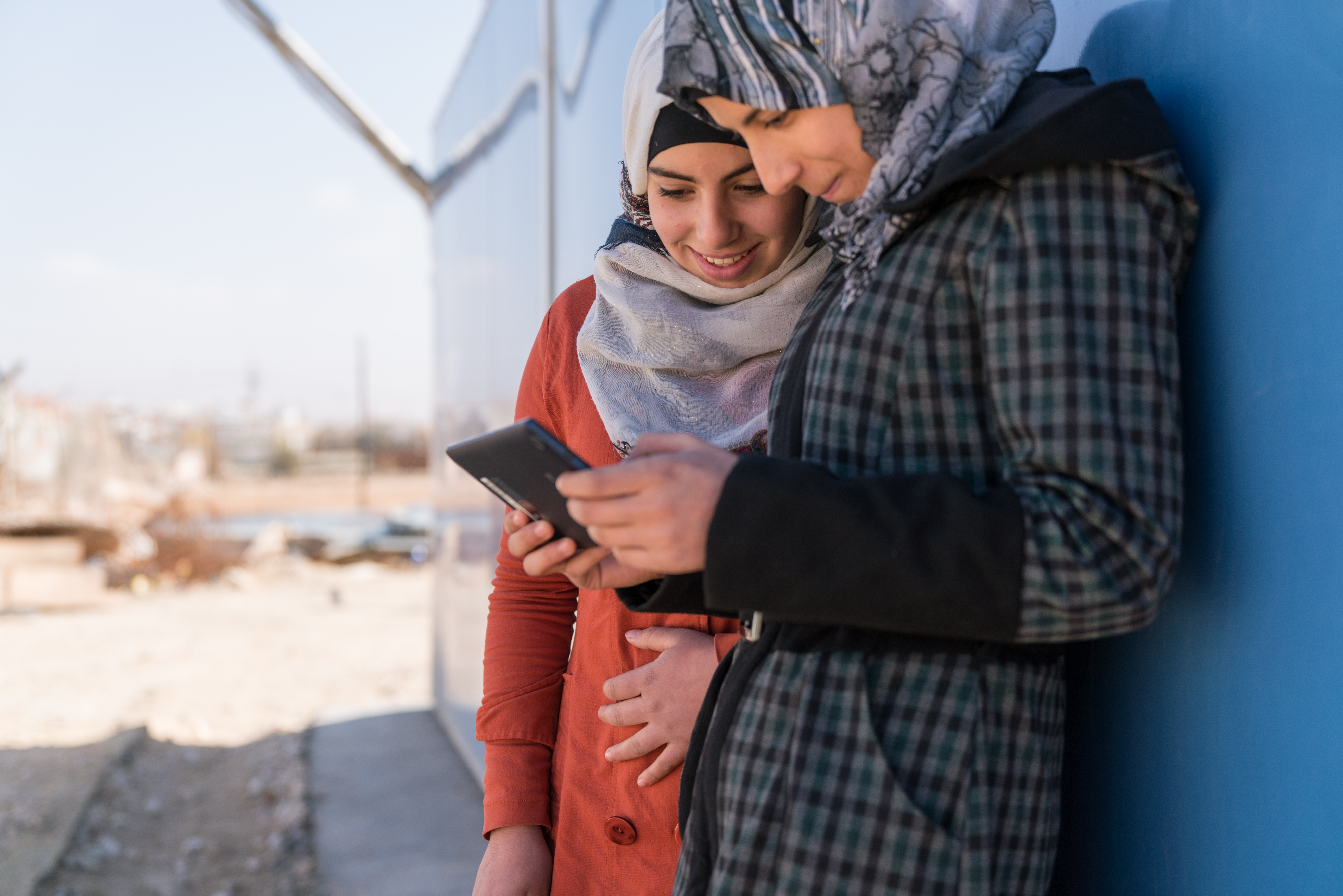 Two girls on a tablet device and leaning against a blue wall.