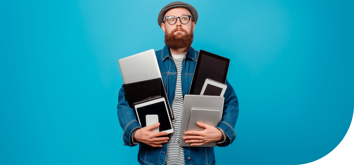A man with a beard, glasses, hat, and denim shirt is holding several electronic devices such as smartphones, laptops, and tablets in his arms. He is standing in front of a neutral blue background and looks concerned and expectant.