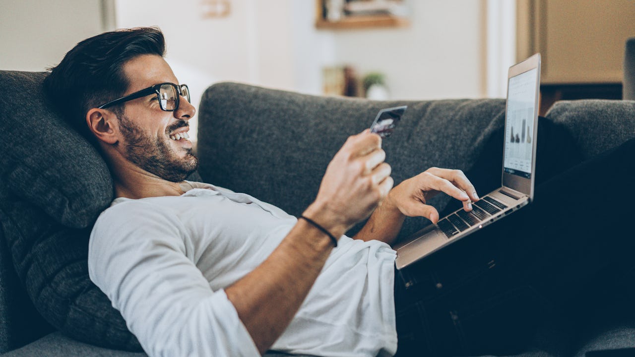 Smiling man lying on the couch and shopping online with credit card and laptop