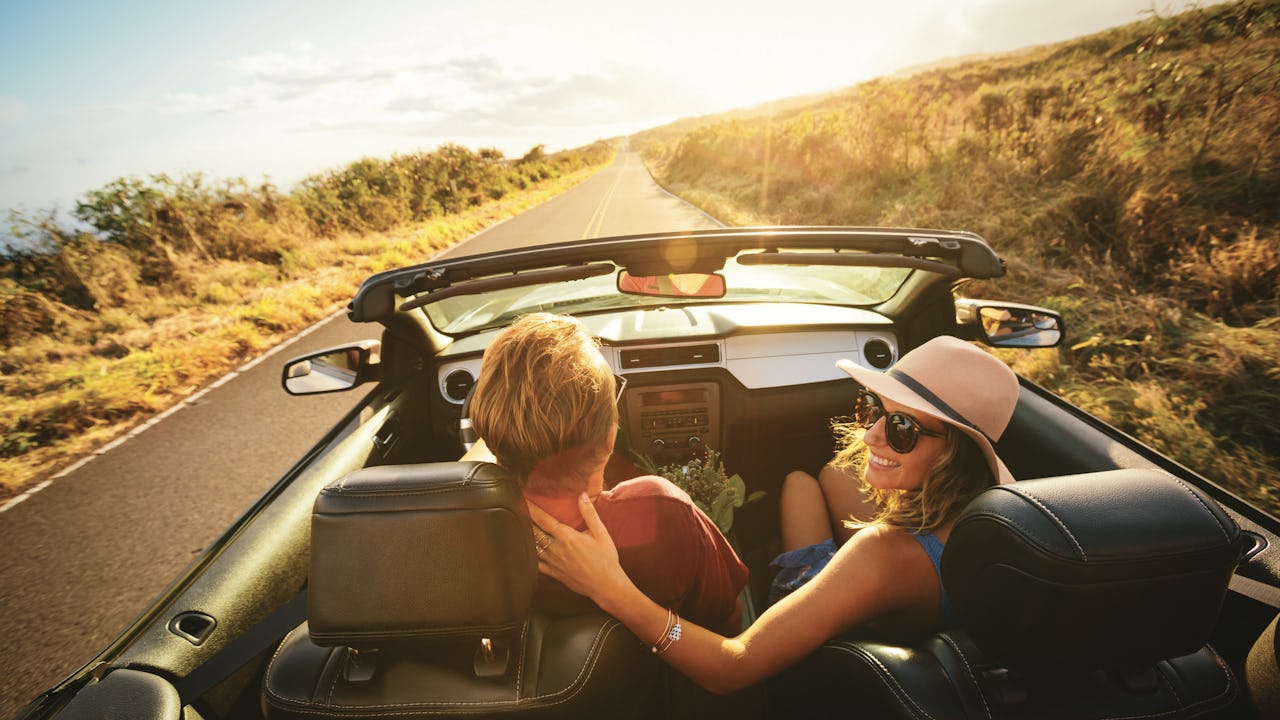 A man and woman driving in a convertible towards the sunset on a country road