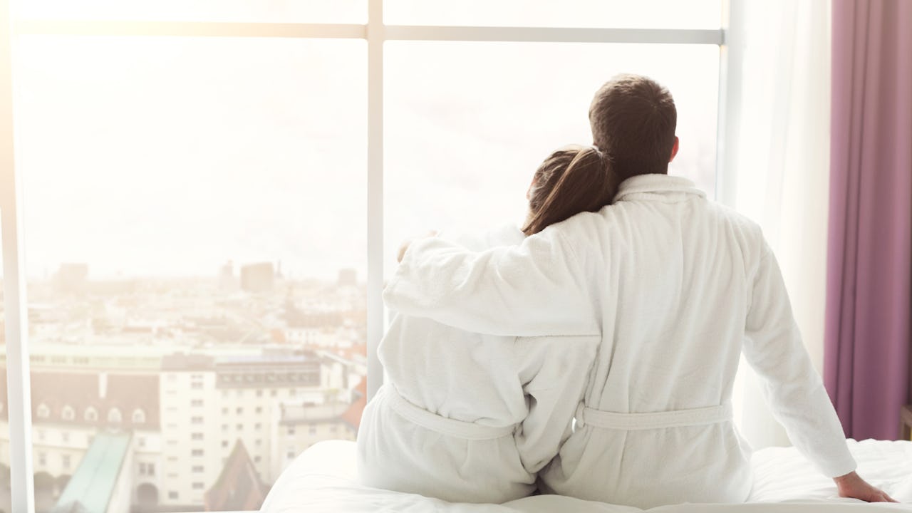 A man with his arm around a woman in their hotel robes, staring out a hotel window