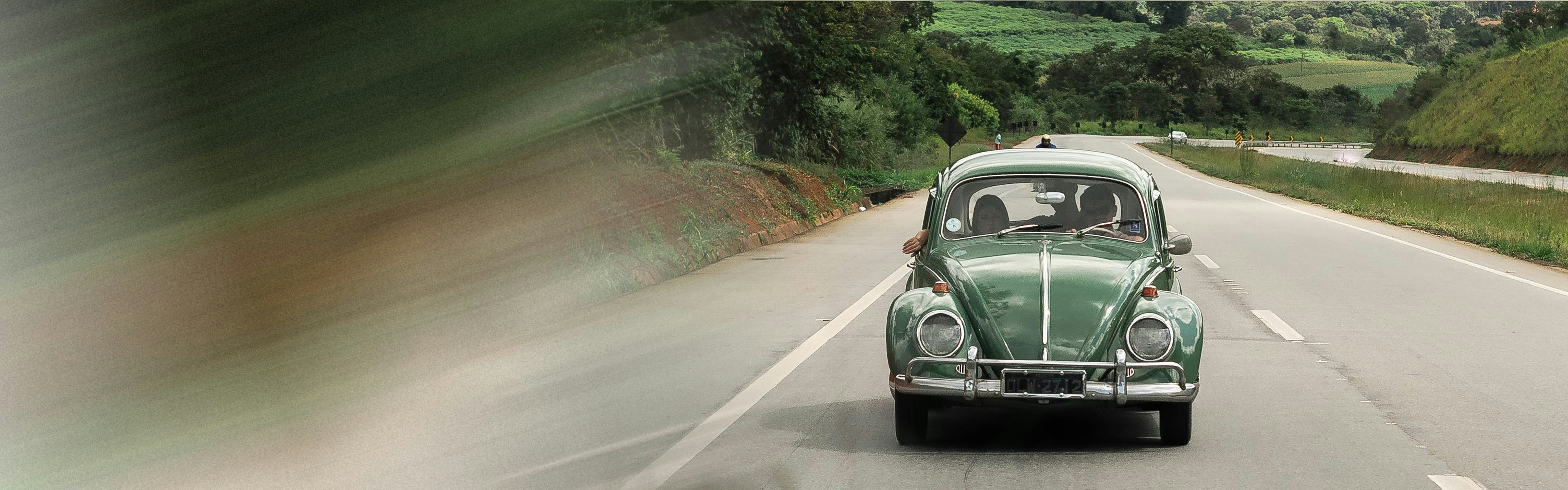Couple in classic green Volkswagen car driving down a highway with forest in background