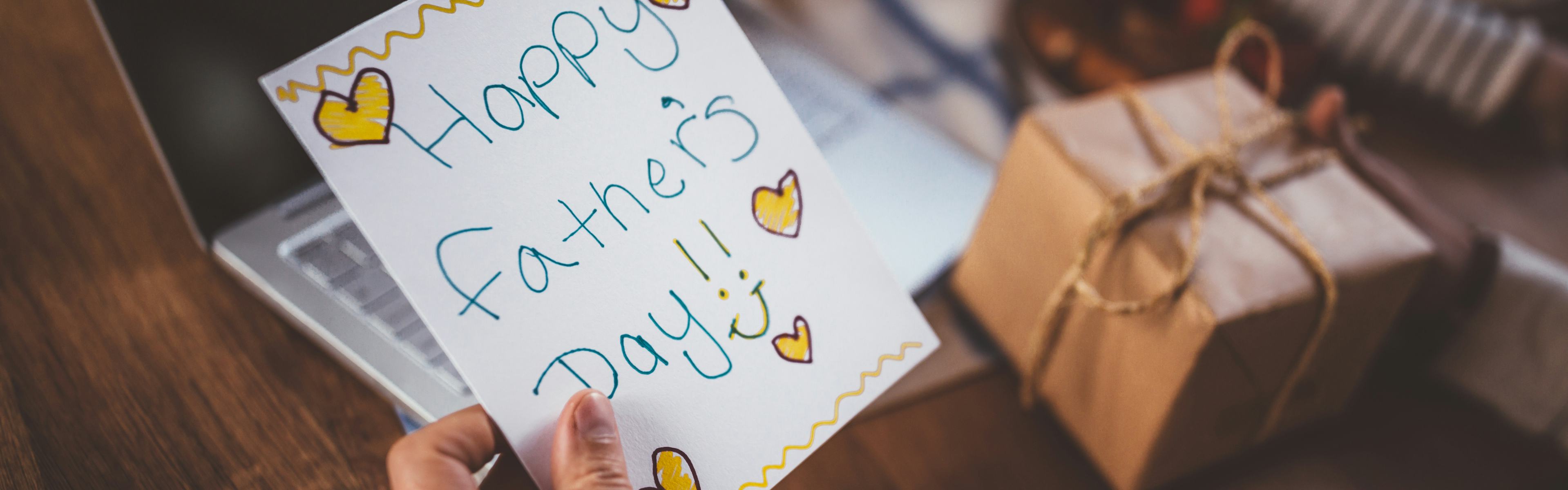 Hand written father's day card being held in front of a table with laptop and wrapped present on it