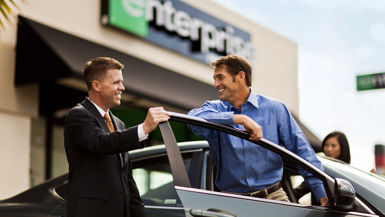 A couple of men smiling beside a car, outside an Enterprise rental office