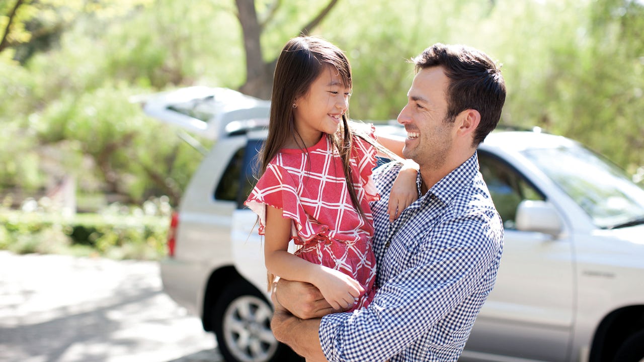 A man holding his daughter, standing in front of a vehicle