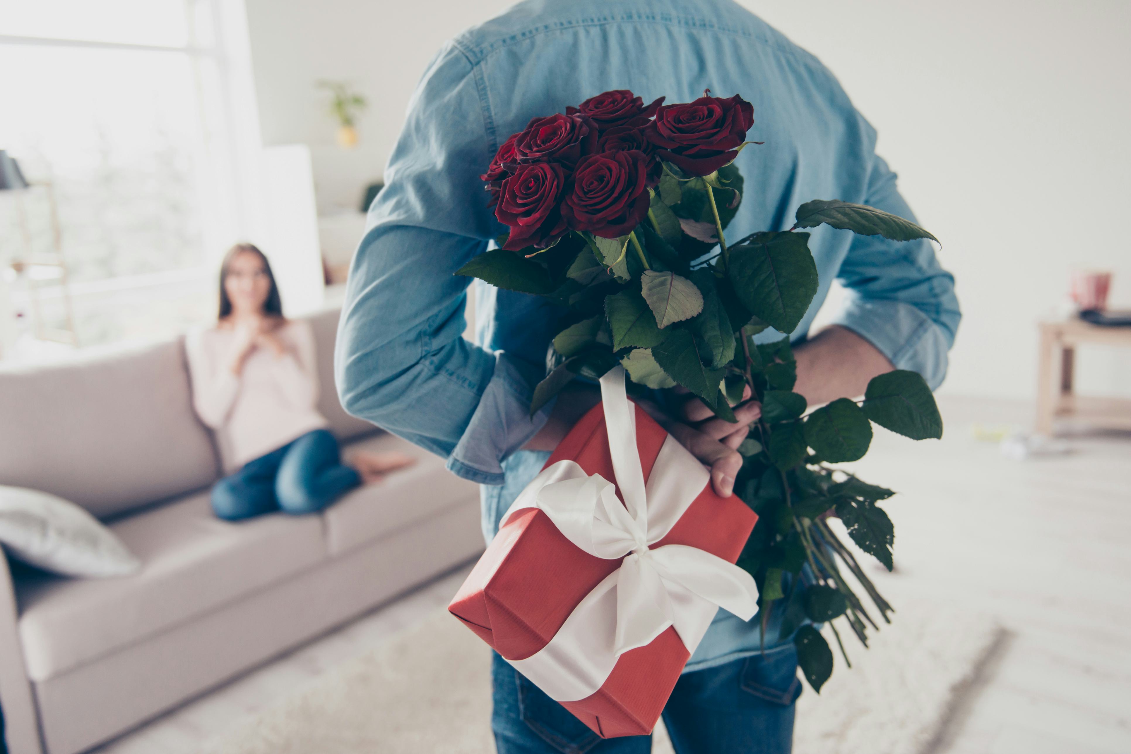 Man holding flowers and a gift behind his back while facing a women sitting on a couch