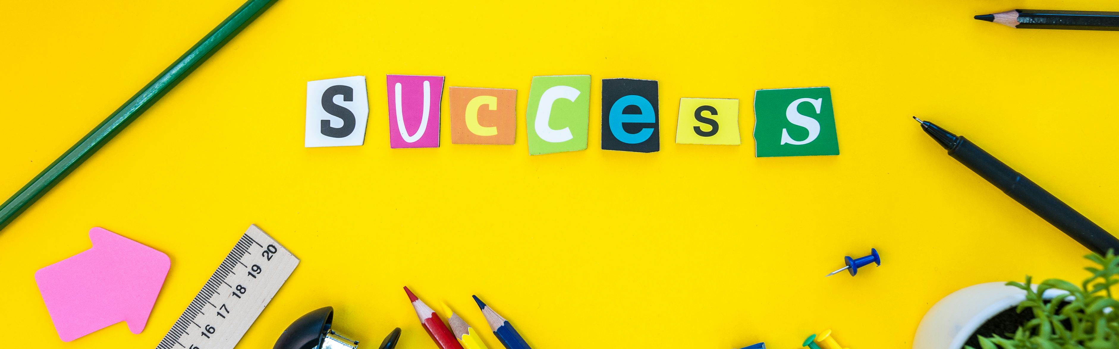 Success - text of carved letters at yellow table background with office or pupil supplies.