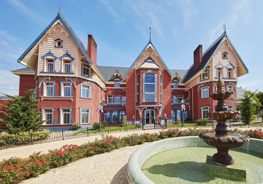 A majestic red brick mansion named Lucy's Mansion, with a fountain, situated near a wonderful theme park.