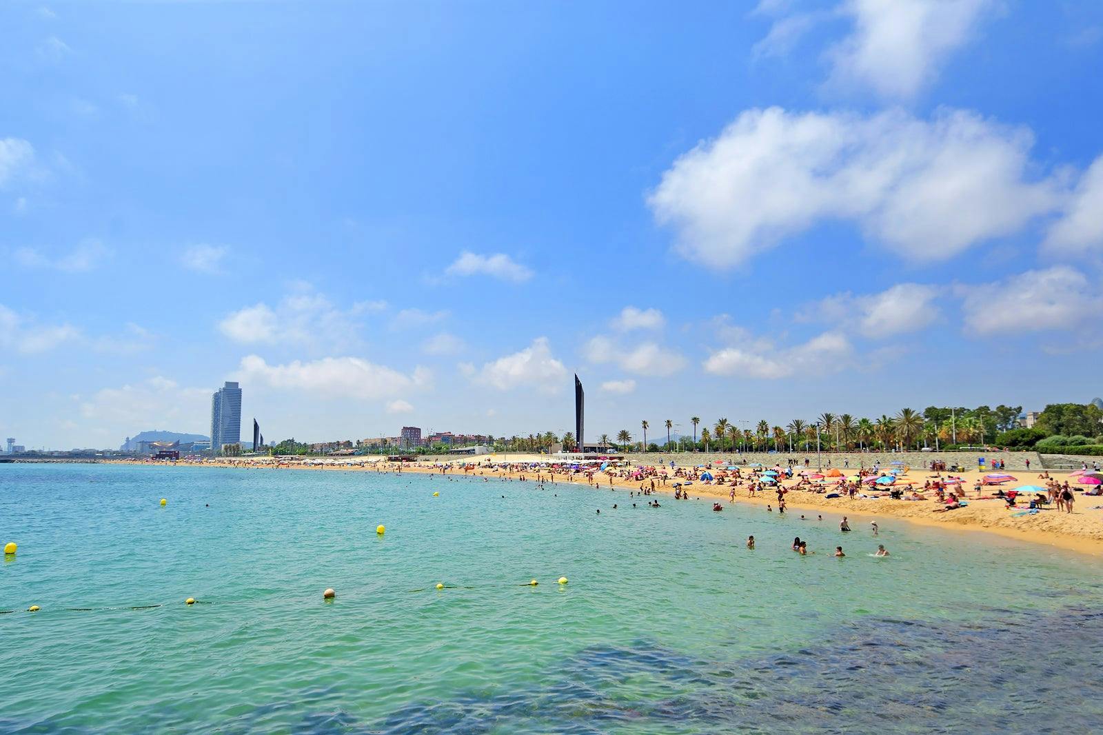 A crowded beach called Bogatell Beach in Barcelona, with numerous people enjoying a swim in the water.