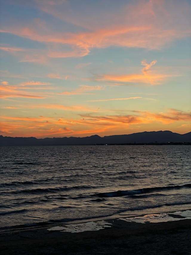 The sun is setting over the ocean with mountains in the distance.