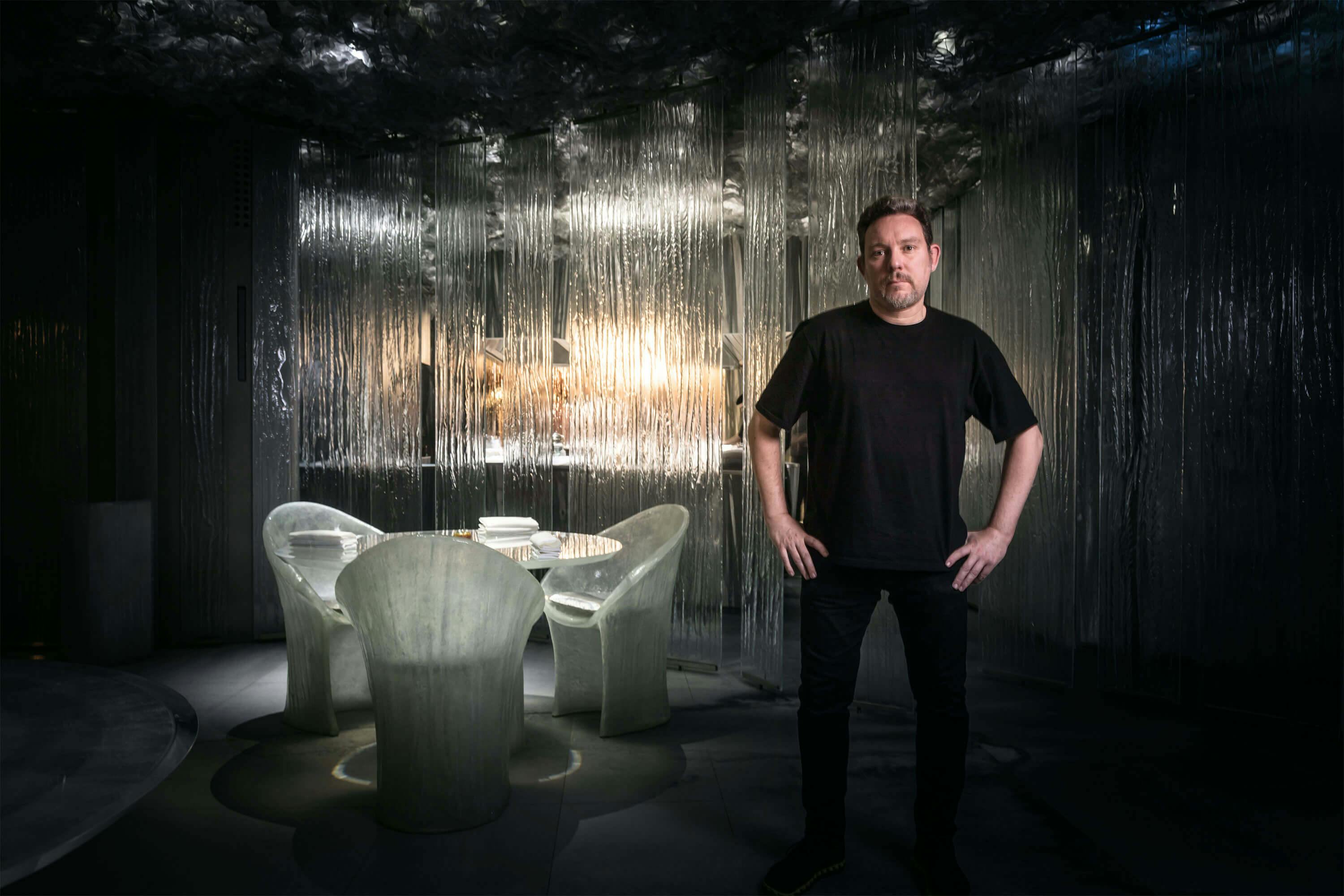 A man, Albert Adrià, stands before a table and chairs at Enigma.