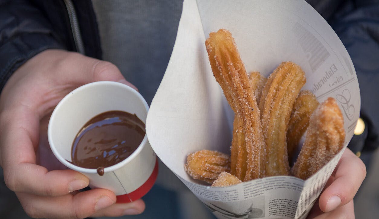 A person holding a cup of churros and a paper, accompanied by a steaming cup of hot chocolate.