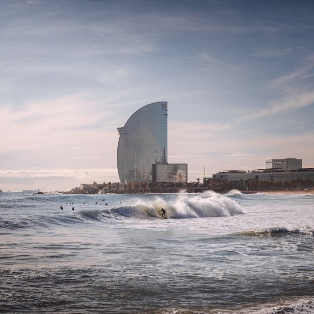  Surfers ride waves with Barcelona skyline in background.