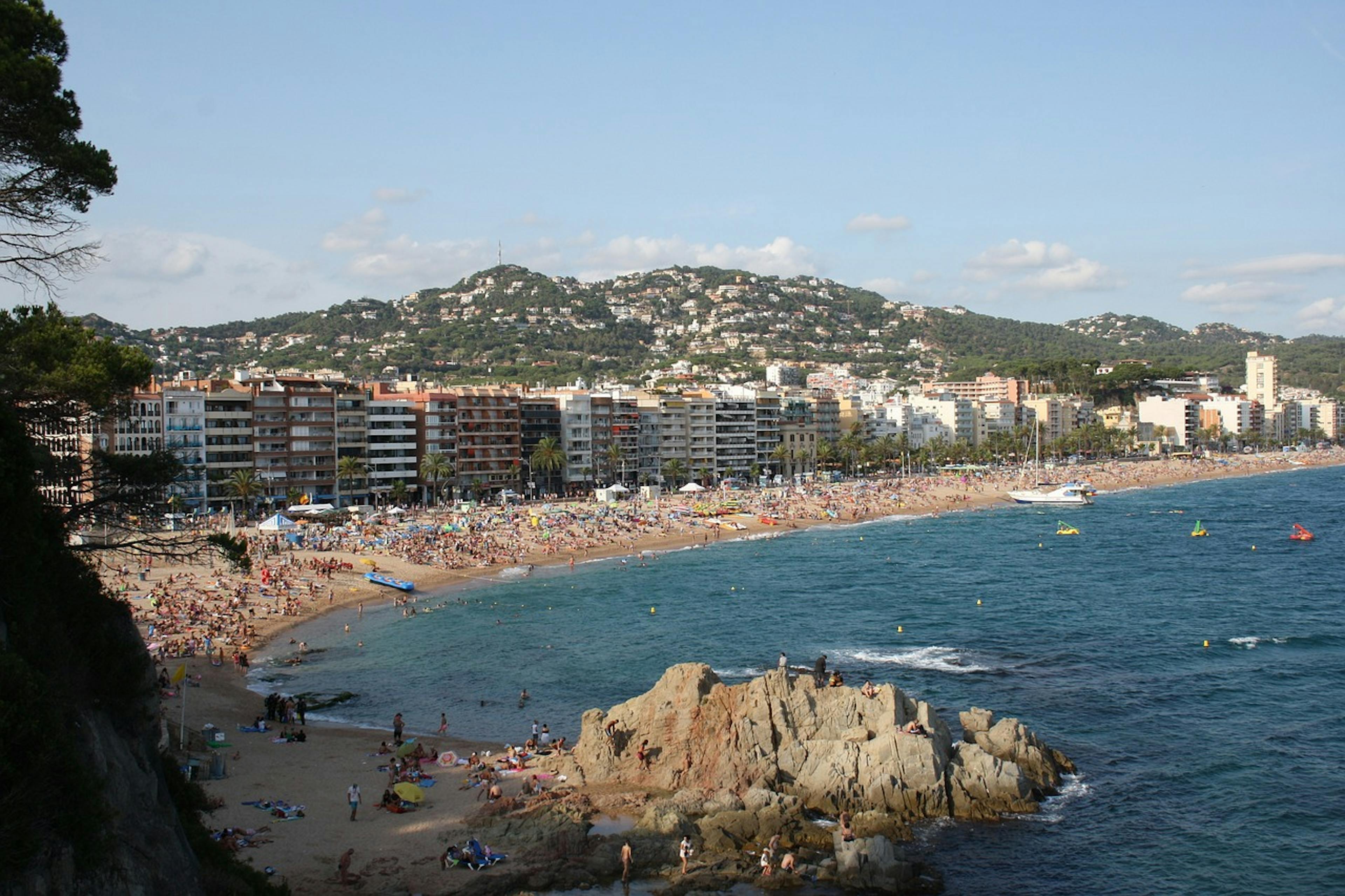 A crowded beach in Lloret de Mar with people swimming and sunbathing under the sun.