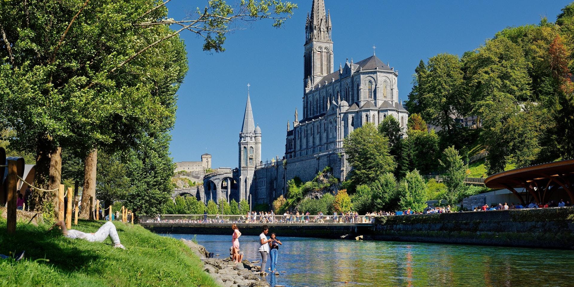 People standing near a castle on the bank of a river, with a basilica in the background.