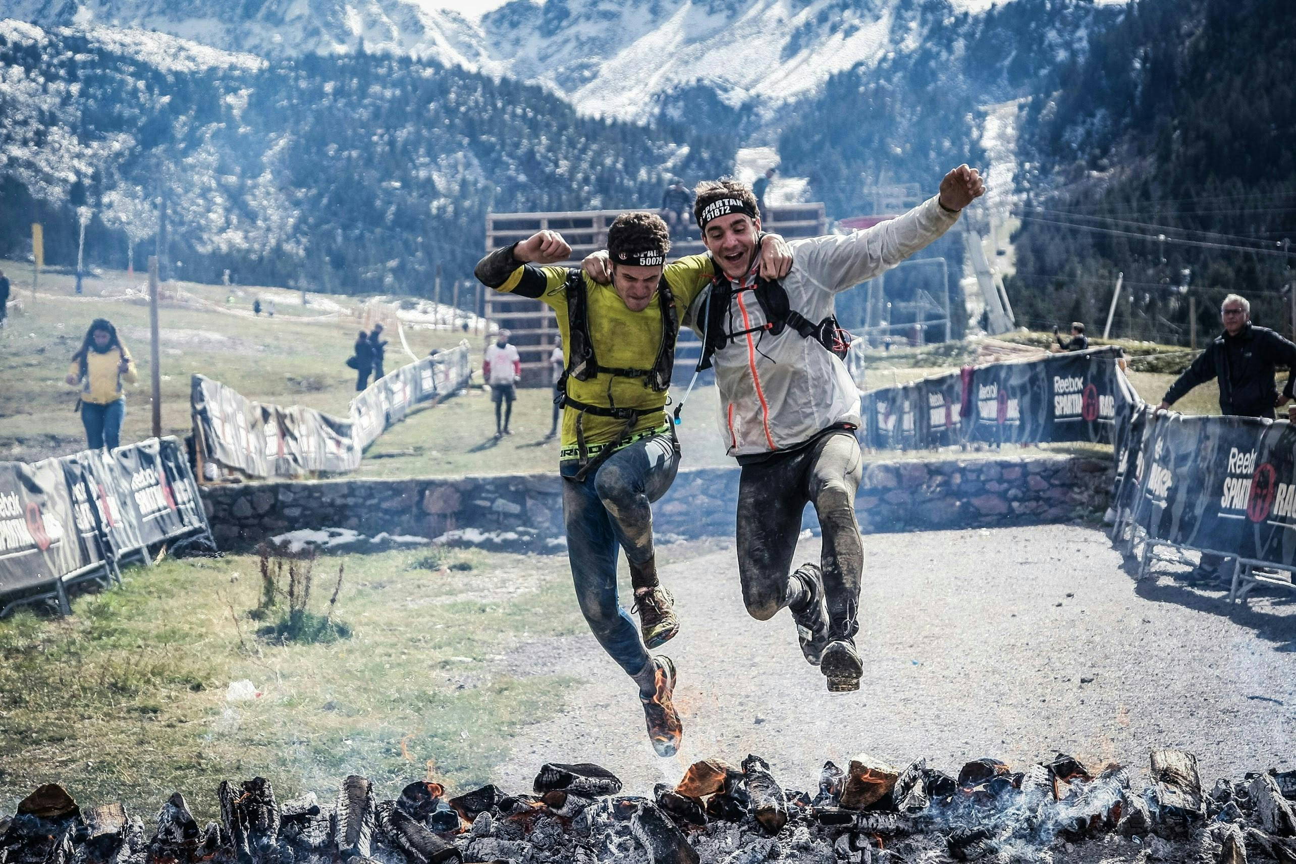 Two men on mountain bikes leaping over a fire pit in the scenic mountains.