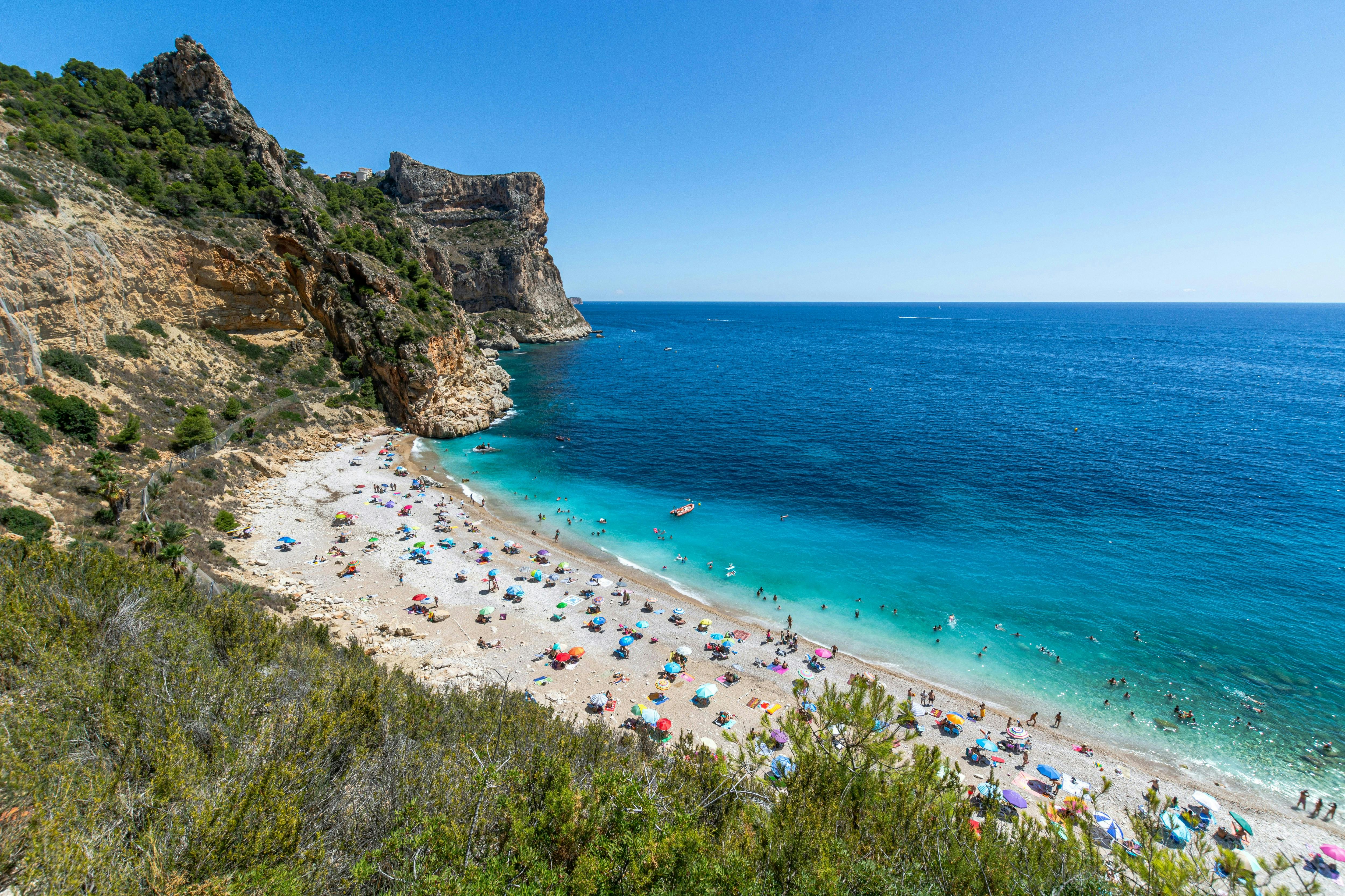 Une plage pleine de monde profitant du soleil et une imposante falaise à Jávea.