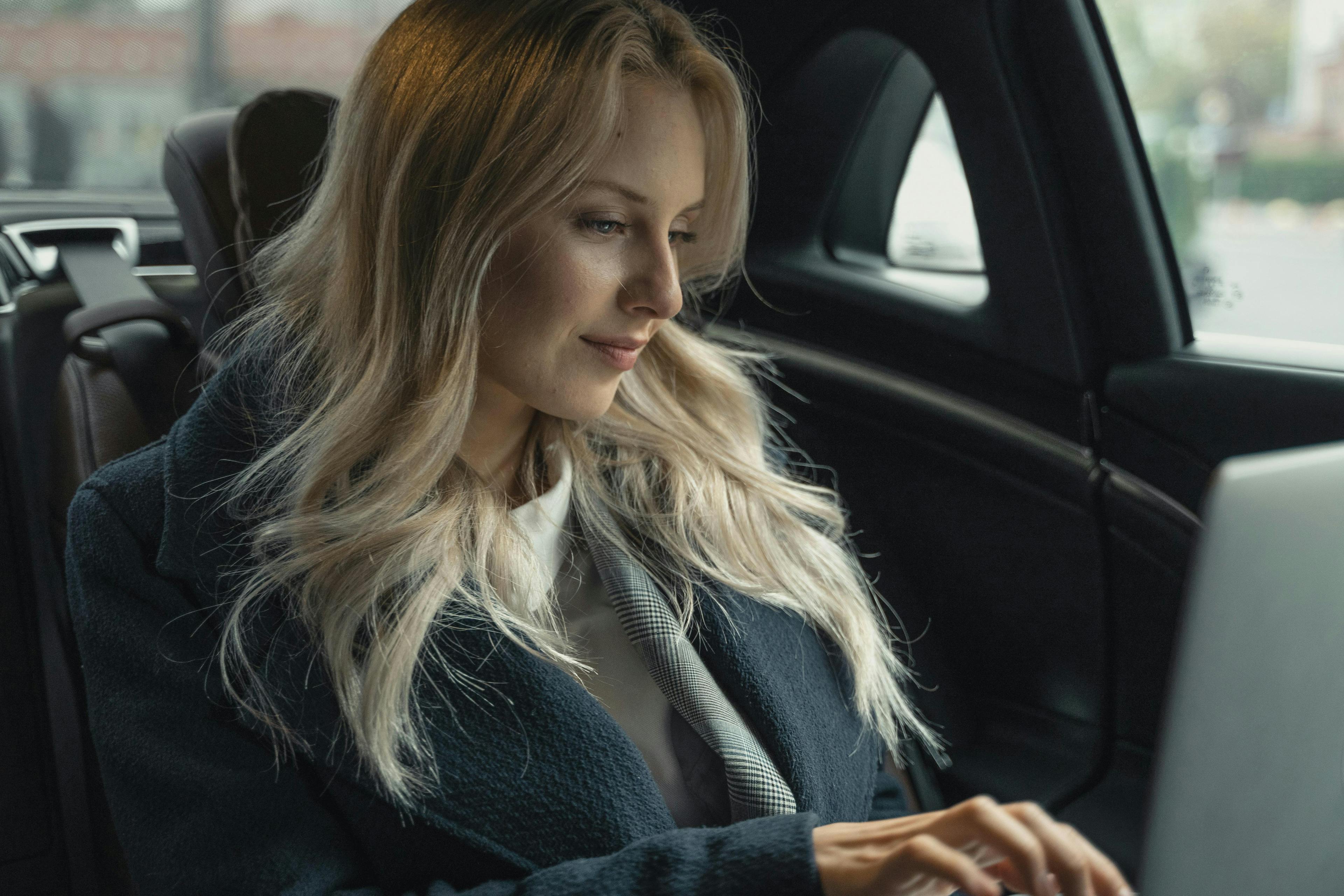 A woman in business attire sits in the back seat of a car, focused on her laptop while traveling for work.