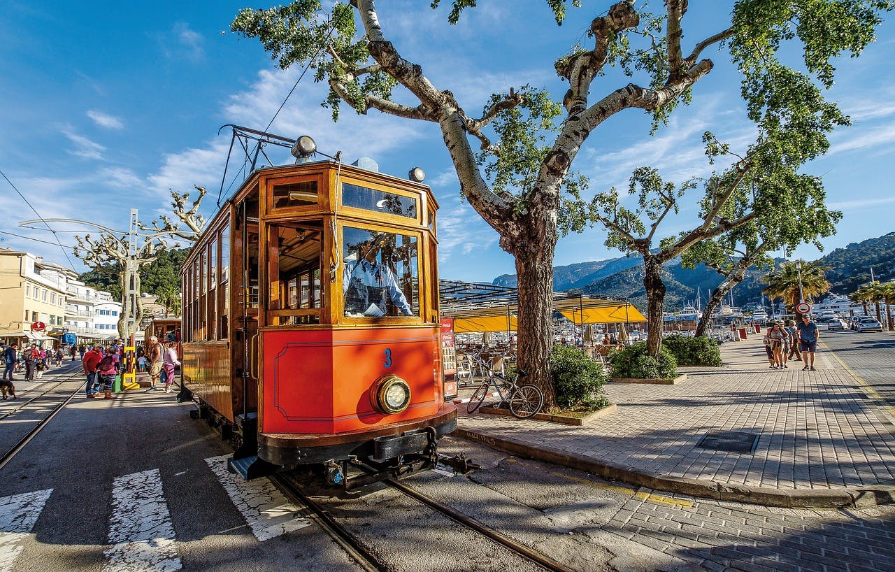 A vibrant red and orange trolley car travels along a picturesque street in Sóller, Mallorca, showcasing local charm.