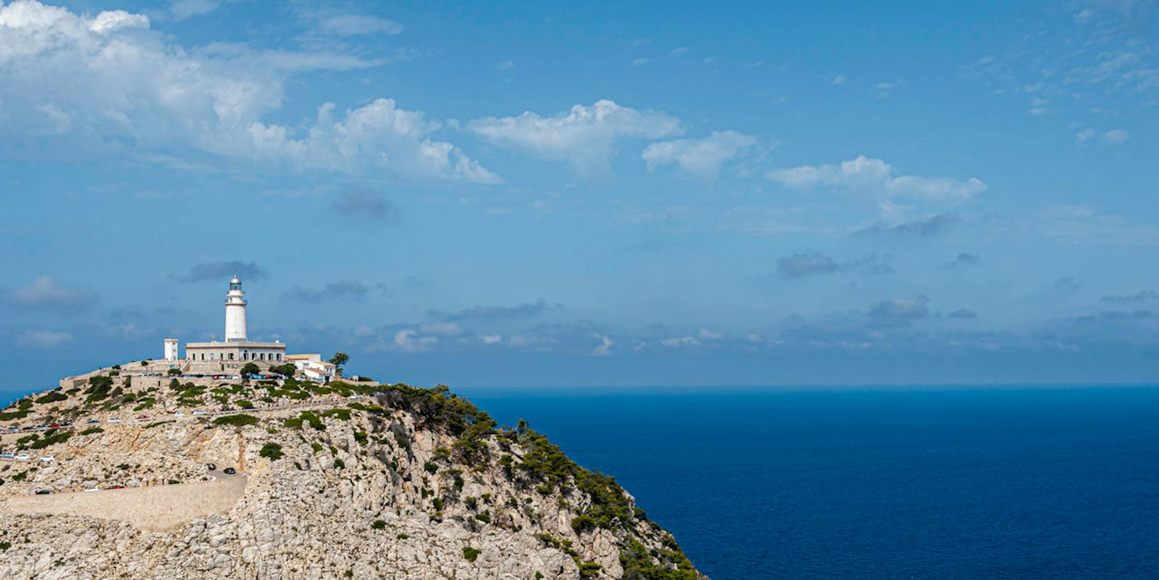 A lighthouse stands on a rocky cliff in Port de Pollença, overlooking the vast ocean under a clear blue sky.