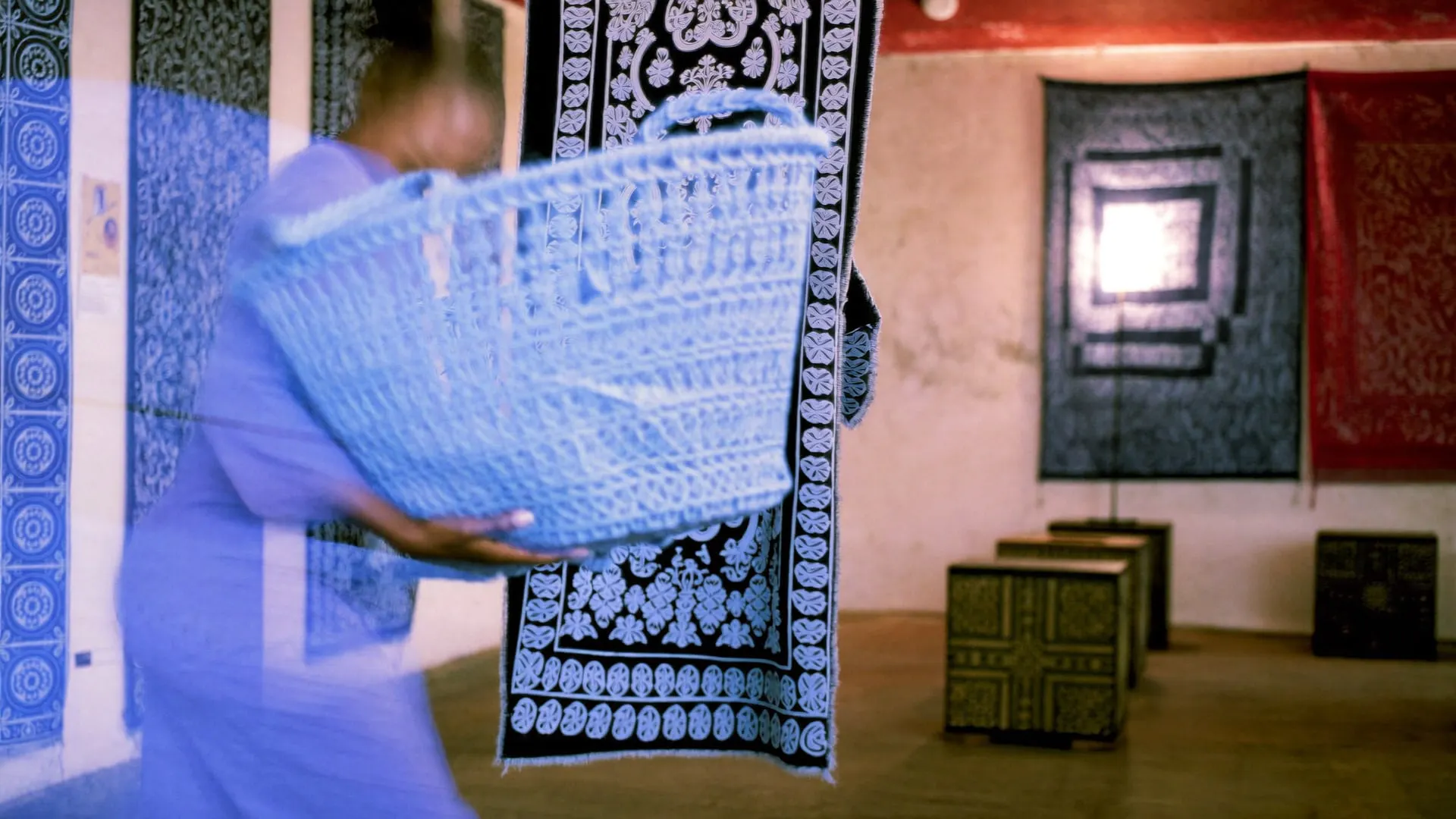 A young woman carrying a woven basket through a gallery of hanging indigo textiles, blurred by a slow shutter.