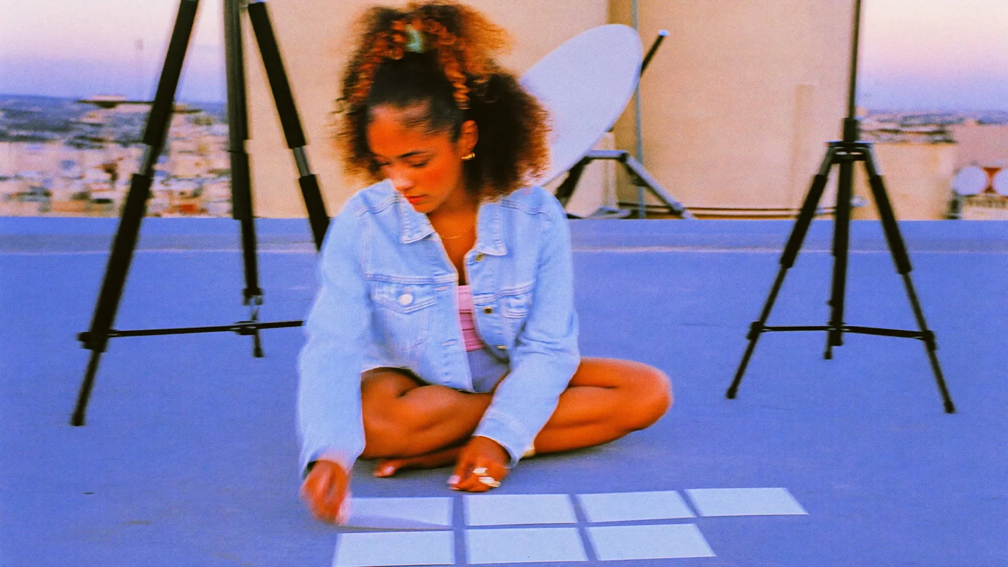 A young woman sitting cross legged on a Cairo rooftop at dusk, laying out blank shot cards between tripods