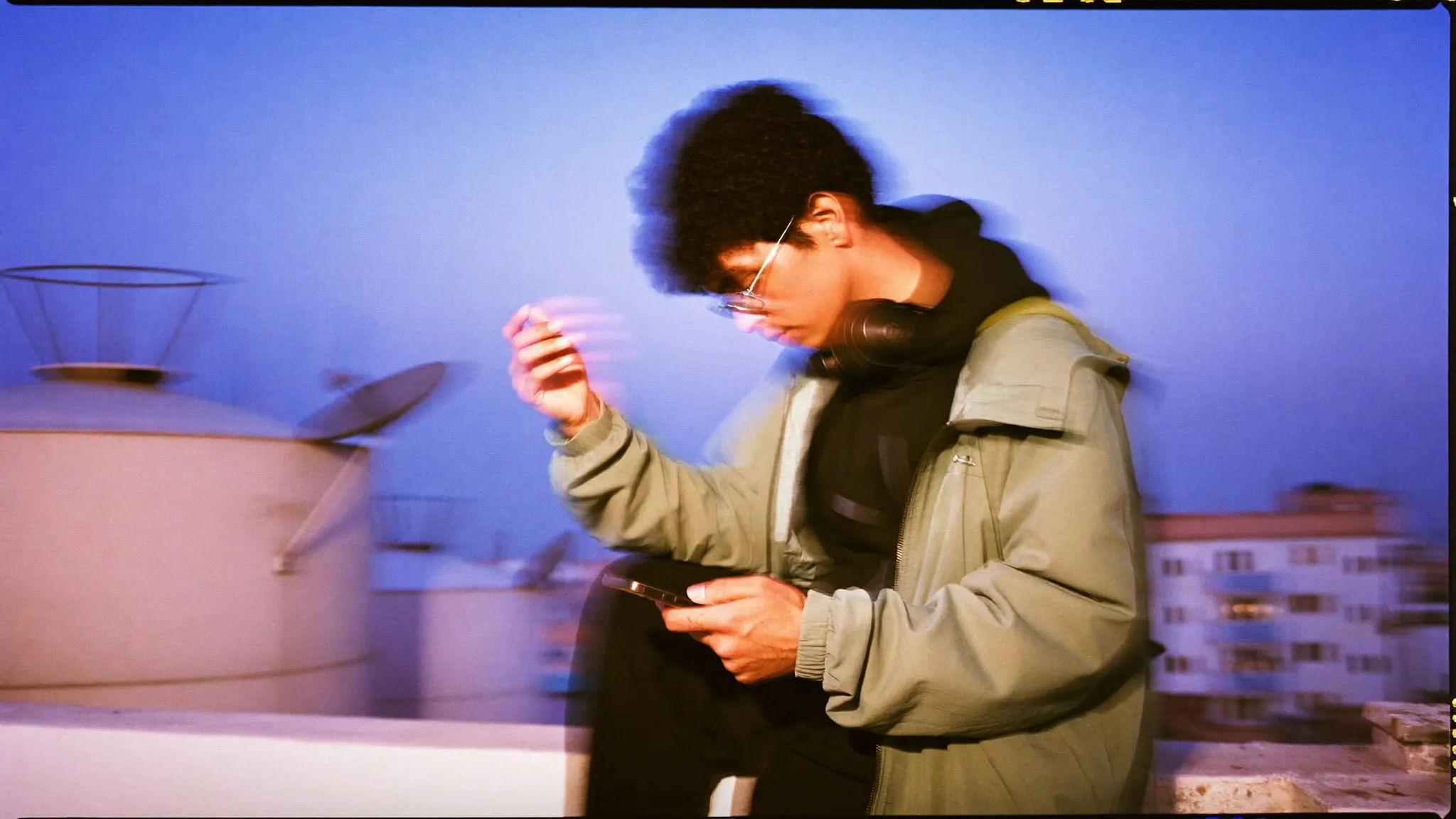 A young man with glasses on a Cairo rooftop at dusk, checking a phone held at arm's length