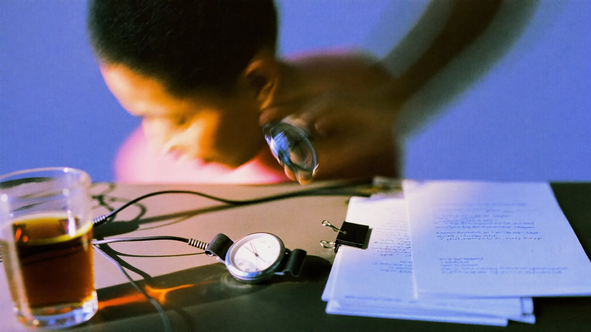 A hand reaching for a stopwatch on a desk with cables, clipped papers and a glass of tea