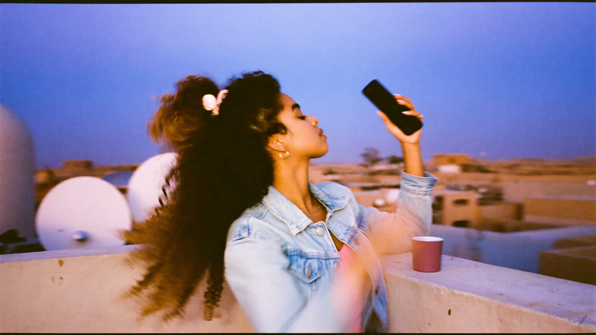 A young woman with curly hair holding her phone up on a Cairo rooftop full of satellite dishes