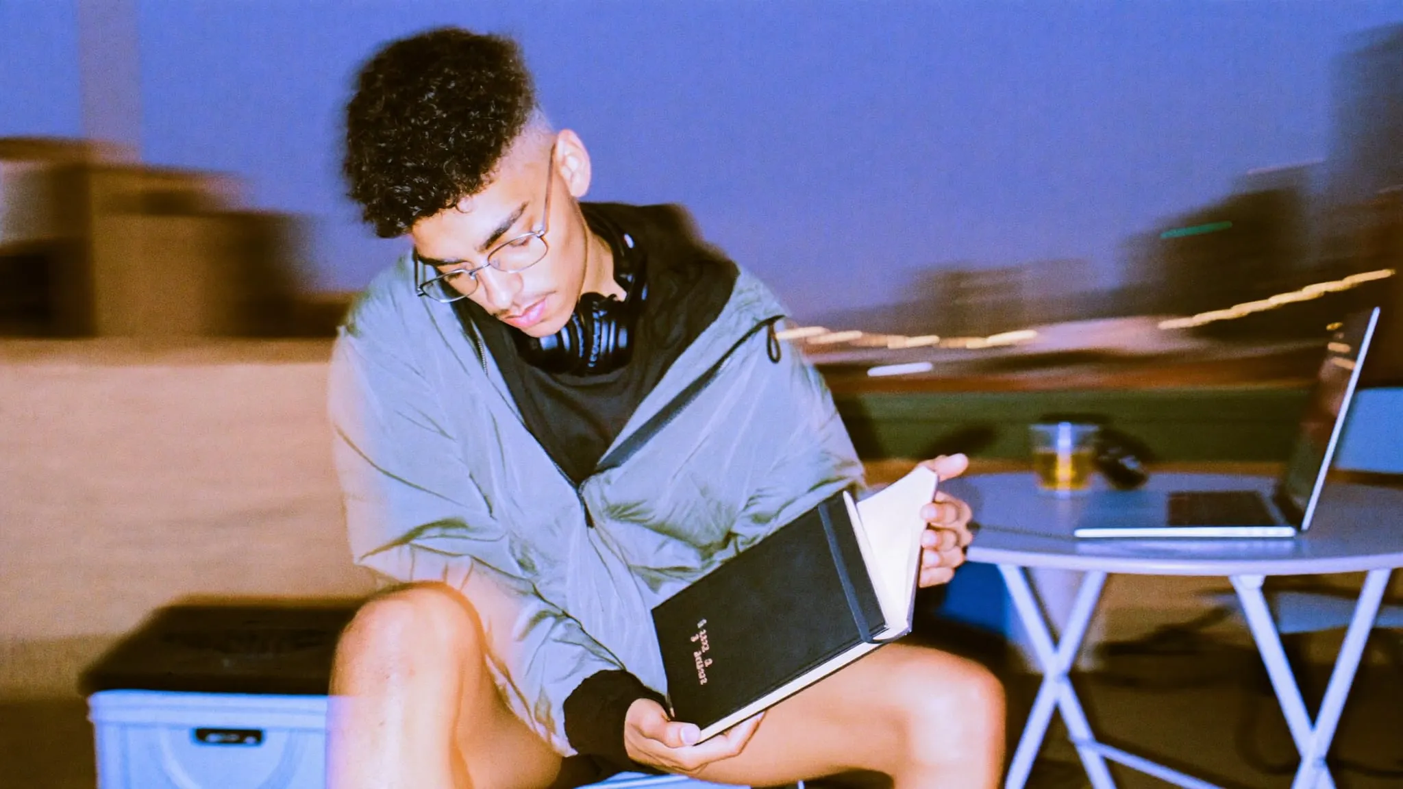 A young man in glasses on a rooftop at night, pen at his lips, holding a black notebook