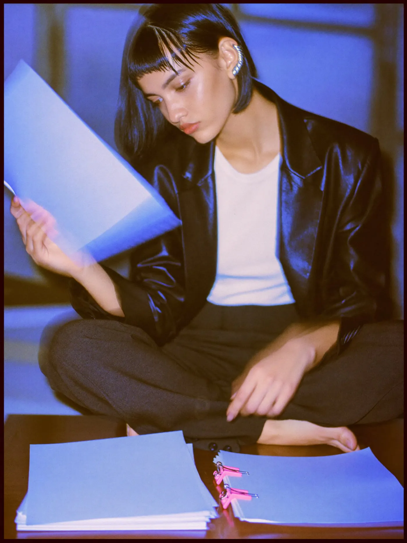 A young woman with a black bob sitting cross legged on a desk, checking a sheet before two paper stacks