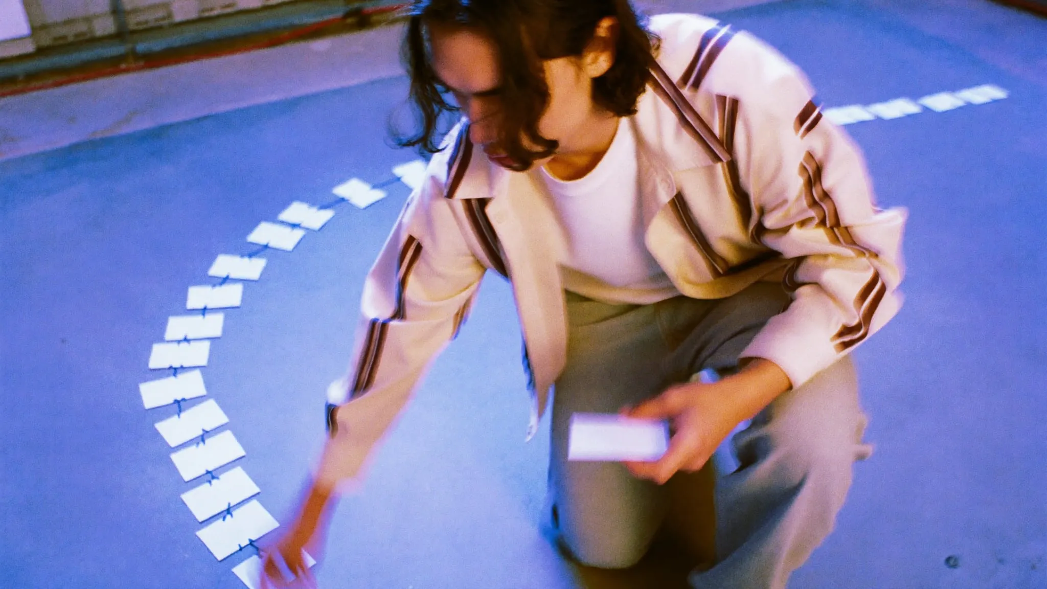 A young man with long wavy hair crouching on a studio floor, placing blank cards in taped rows