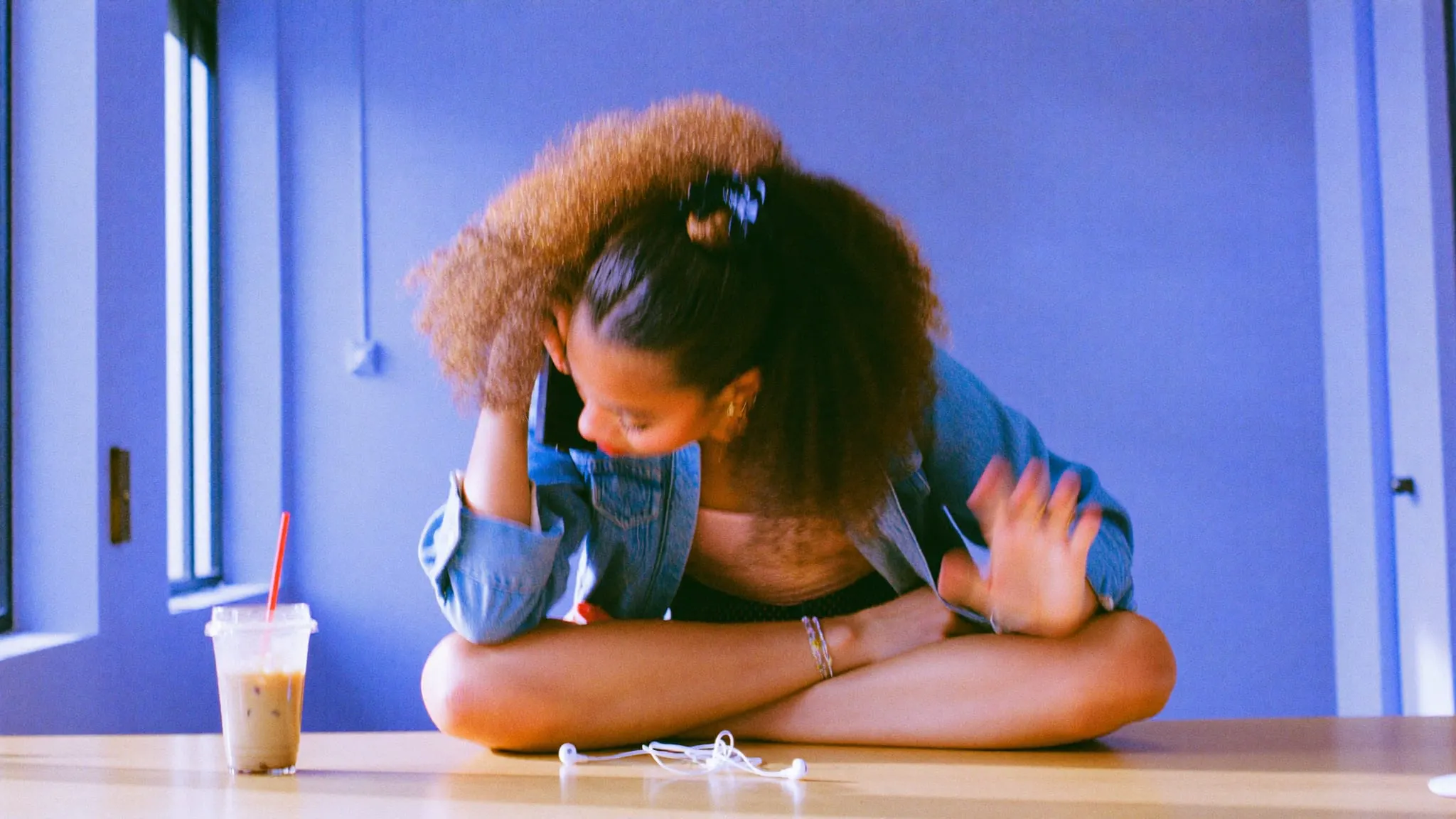 A young woman with curly hair sits cross legged on a desk, on a phone call with a customer