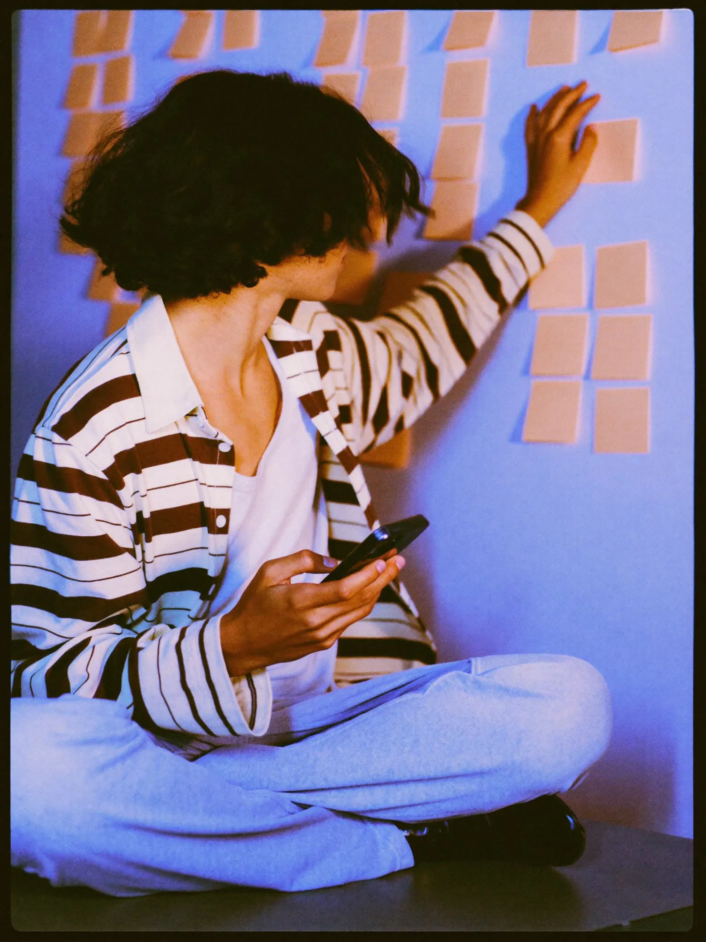 A young man with long wavy hair holds his phone and sticks notes on a wall of grouped sticky notes