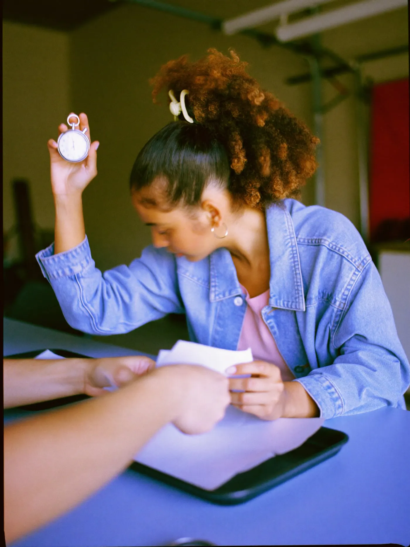 A young woman with curly hair holding up a stopwatch while a colleague moves papers between trays