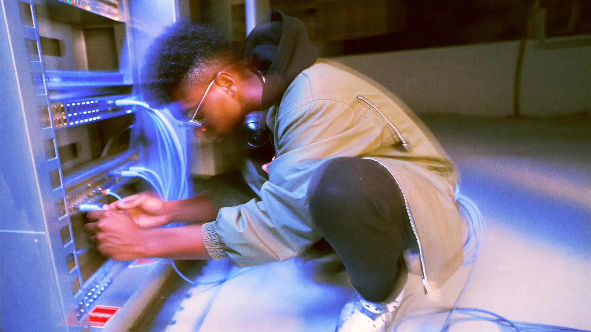 A young man in glasses and a green windbreaker crouches by a network rack, plugging in a blue cable