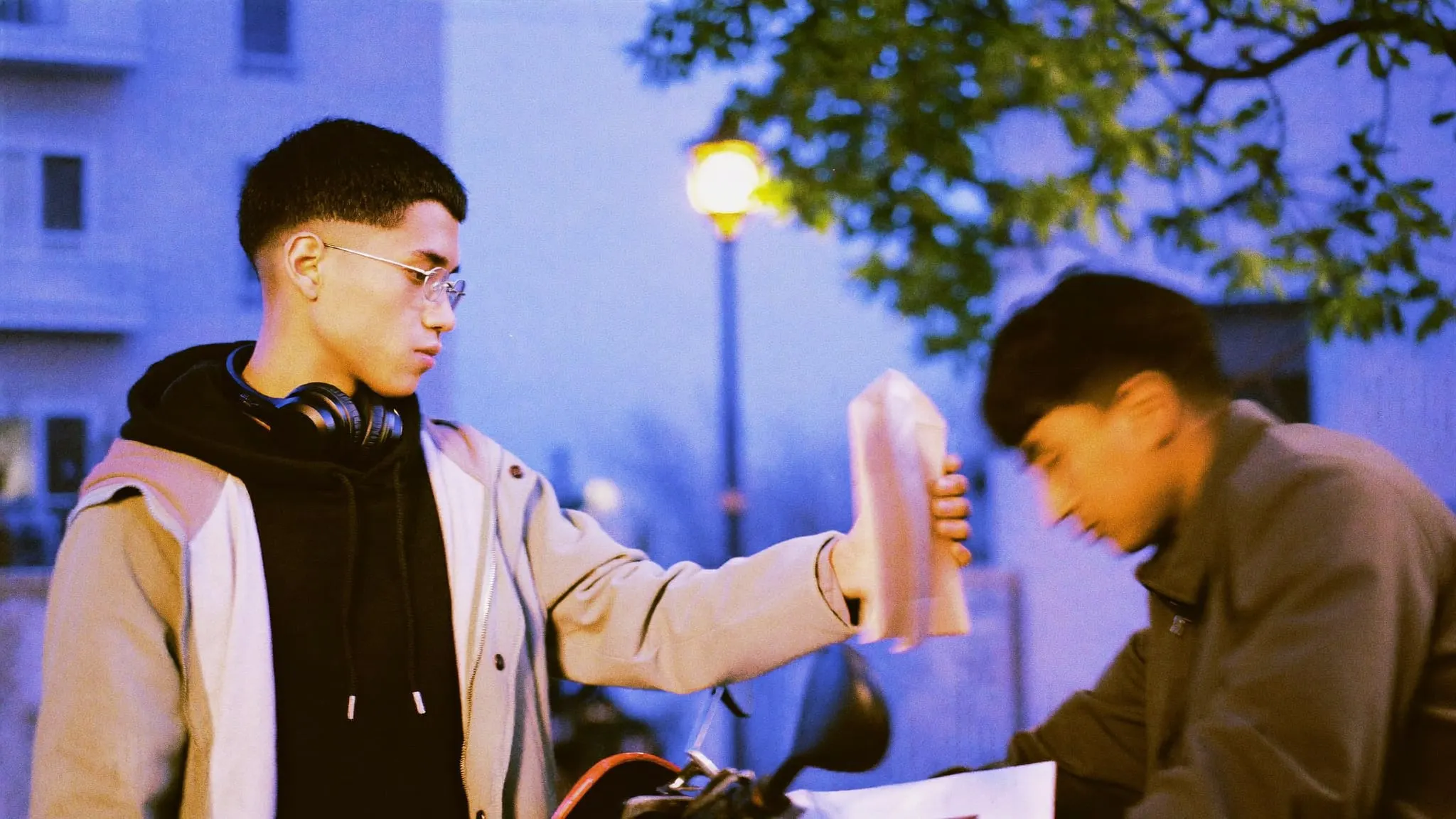 A young man in glasses handing a taped parcel to a delivery rider on a Zamalek street at dusk