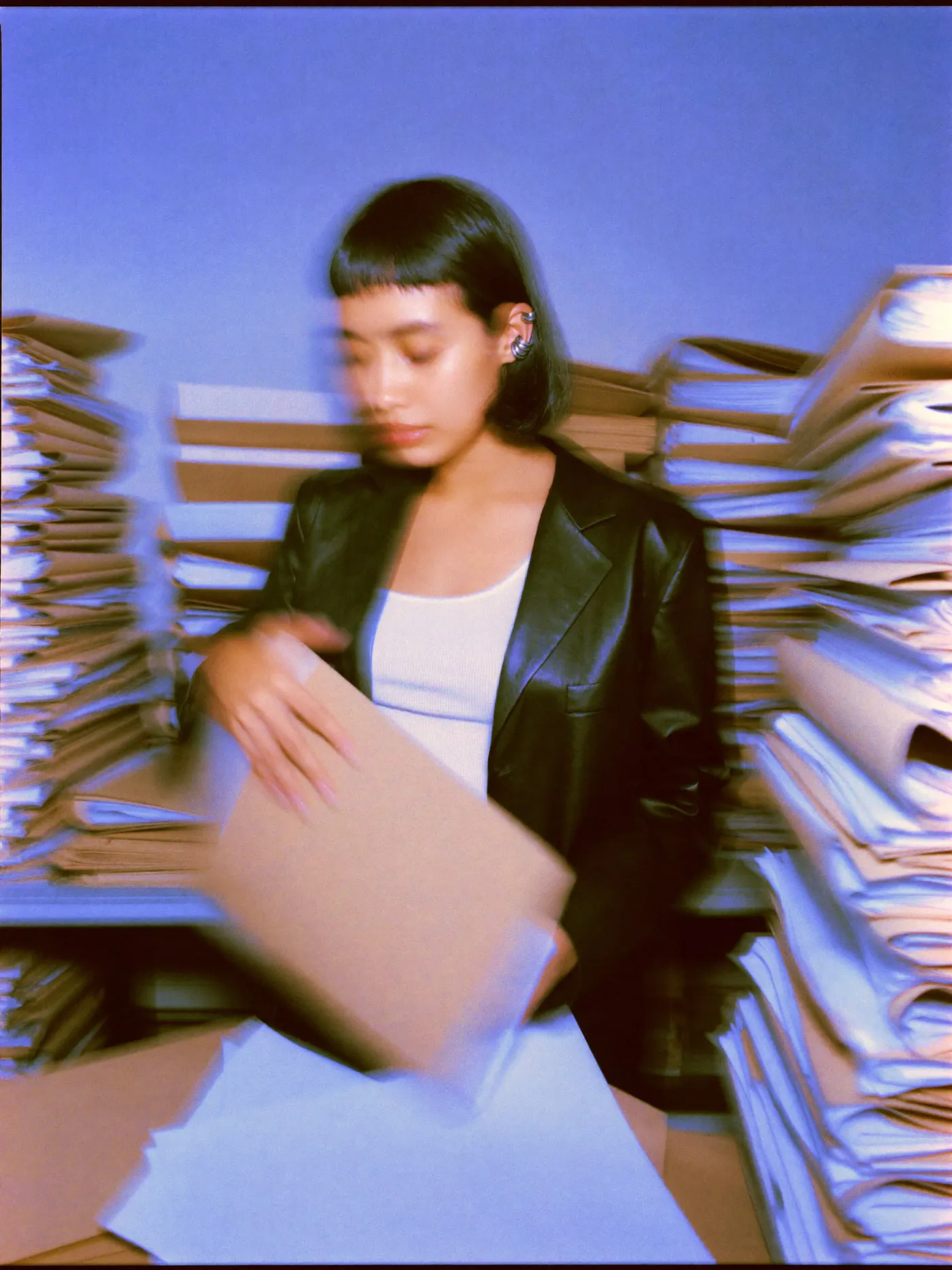 Farida reading a kraft folder in an office stacked with towers of paper files
