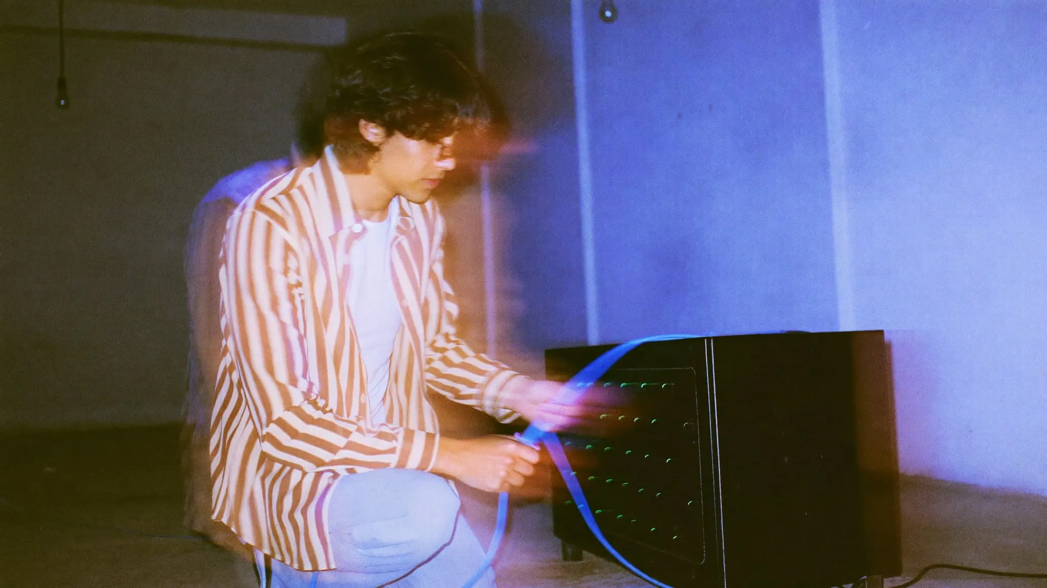 A young man with long wavy hair kneeling in a garage, plugging a blue cable into a server cabinet