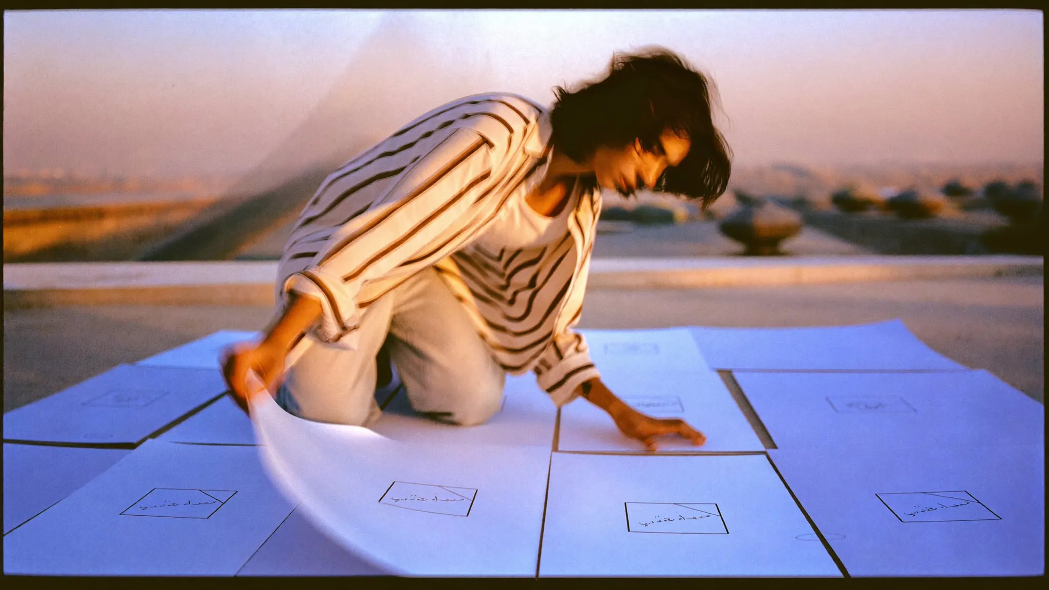 Young man crouching on a Cairo rooftop, laying out sketched website page layouts in a row
