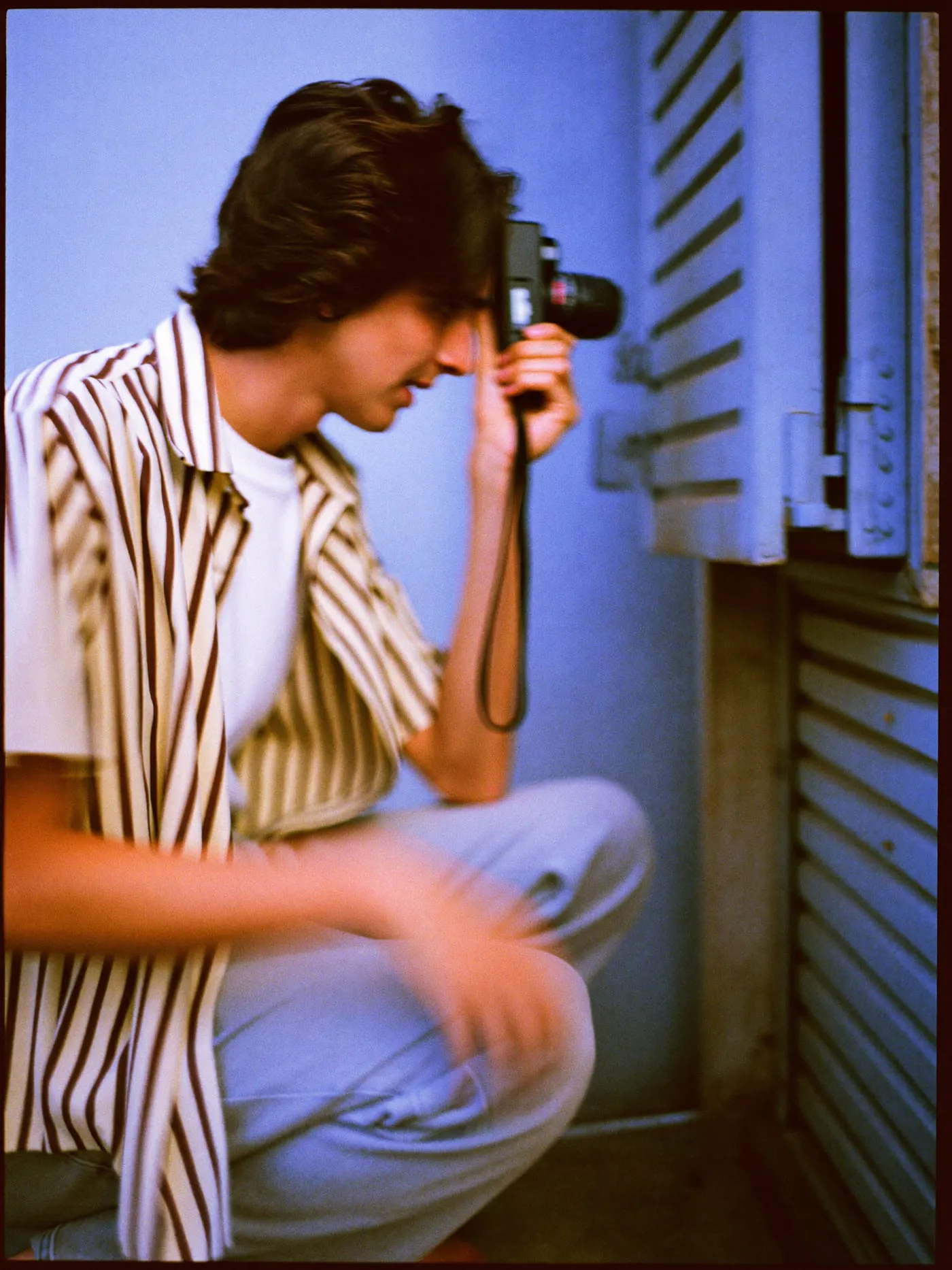 A young man with shoulder length hair crouches on a Cairo pavement, aiming a film camera at an open blue shop door