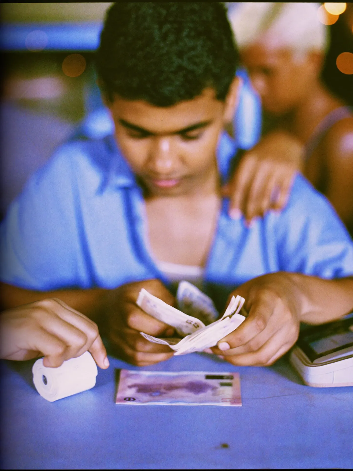Hands counting folded banknotes beside a payment card and a receipt roll on a kiosk counter