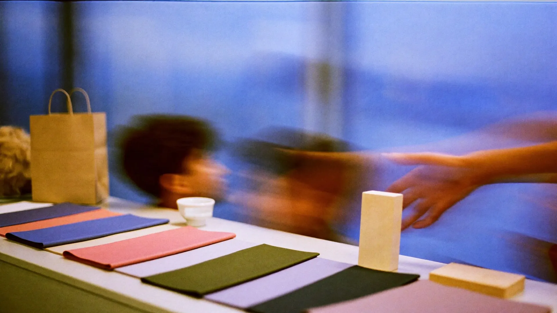 A studio table with fabric swatches, a kraft paper bag, painted blocks and a cup at dusk