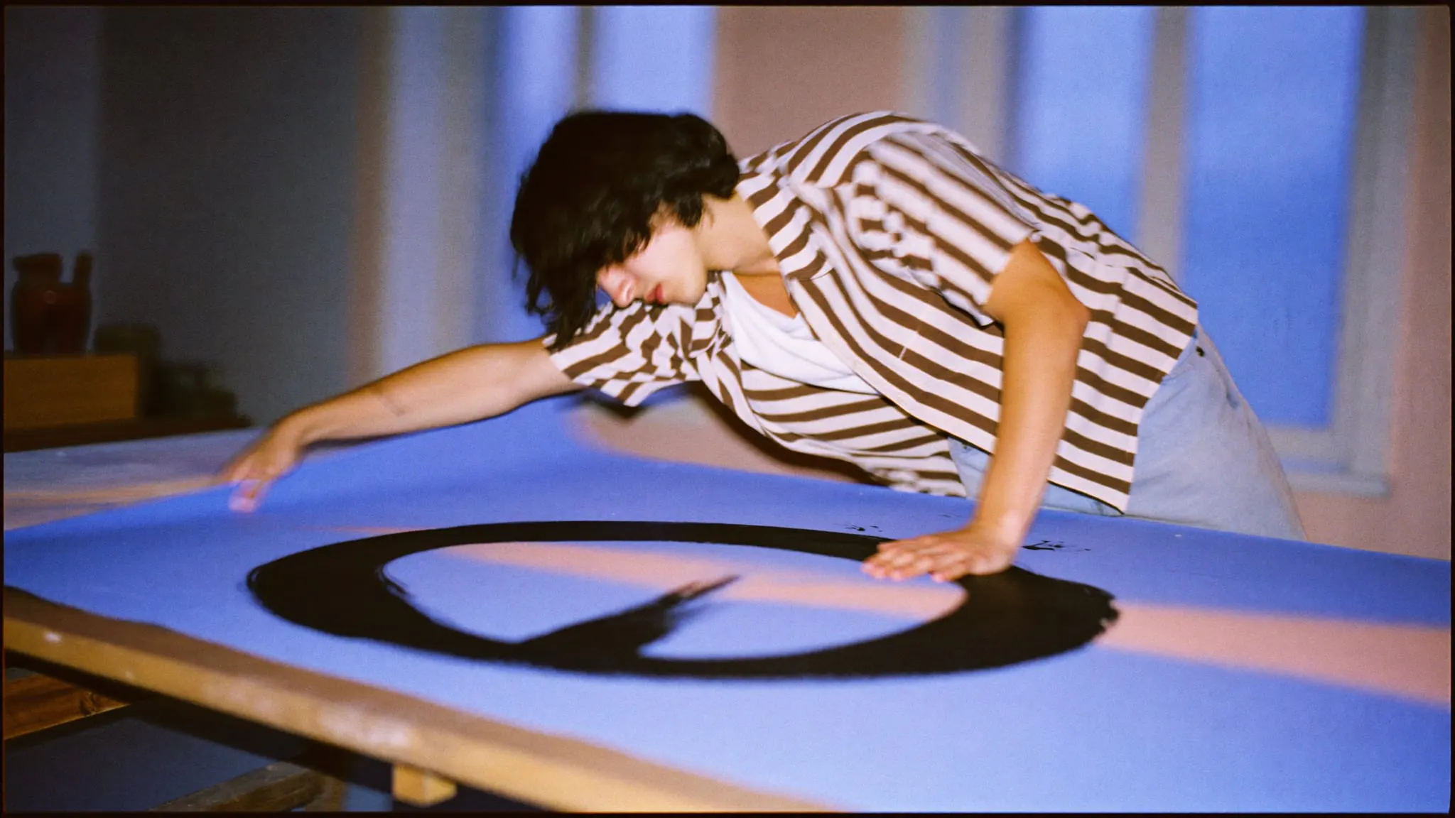 A young designer leaning over a studio table, sliding a large periwinkle sheet of black brush strokes