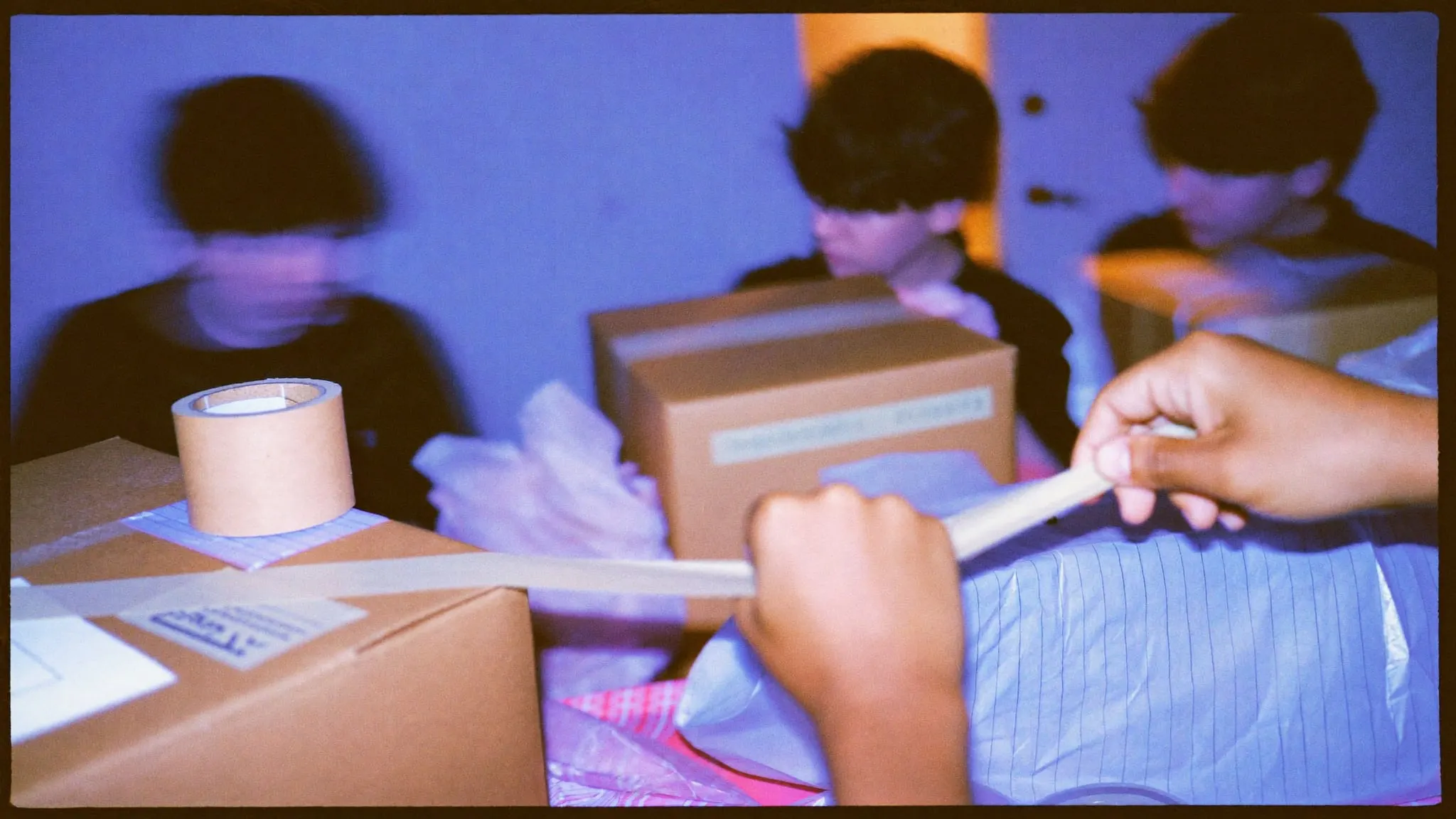 Hands taping a brown mailer box shut on a packing table with tissue paper and tape