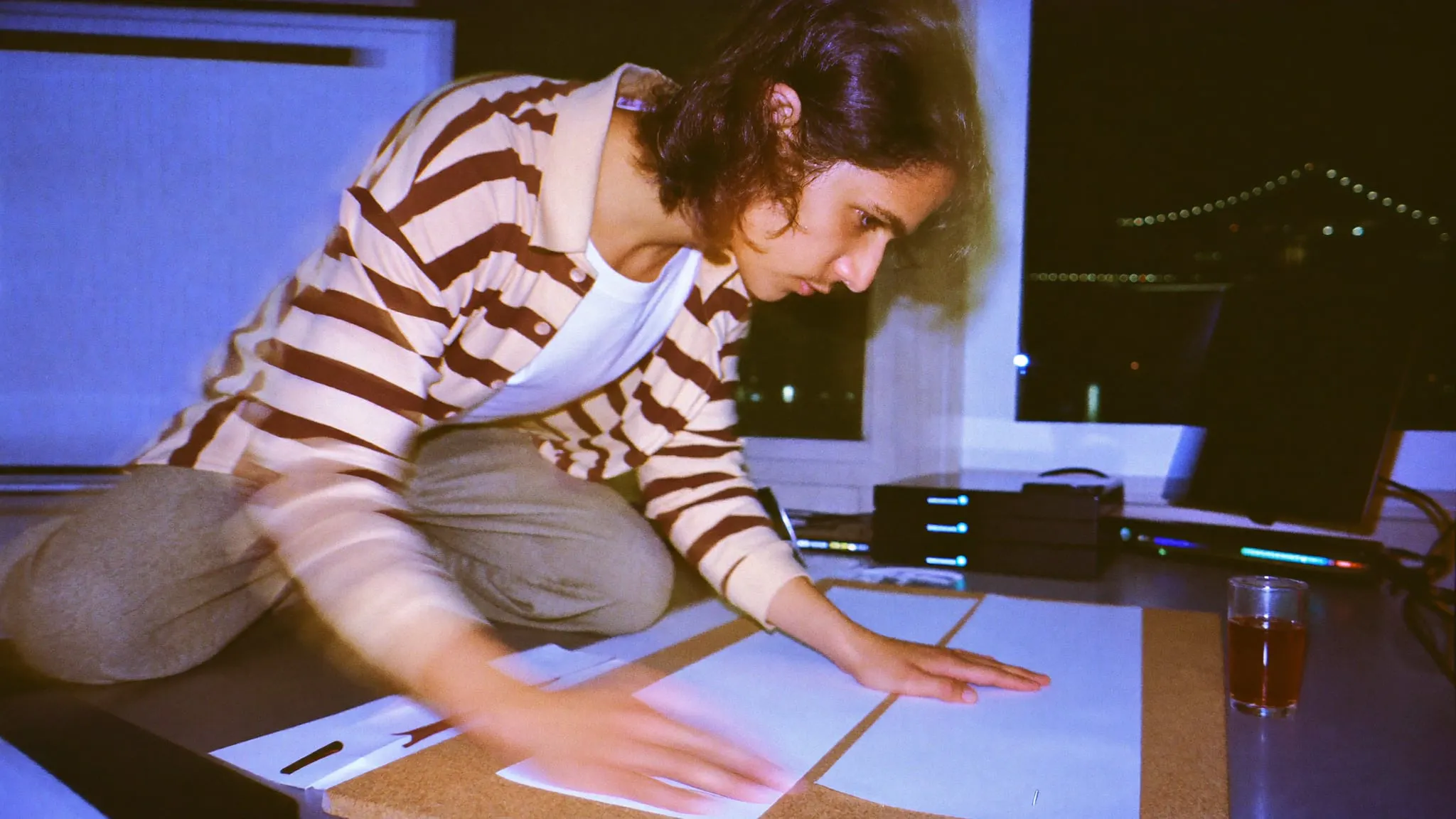A young editor kneeling on a desk at night laying out blank frames, Nile lights through the window