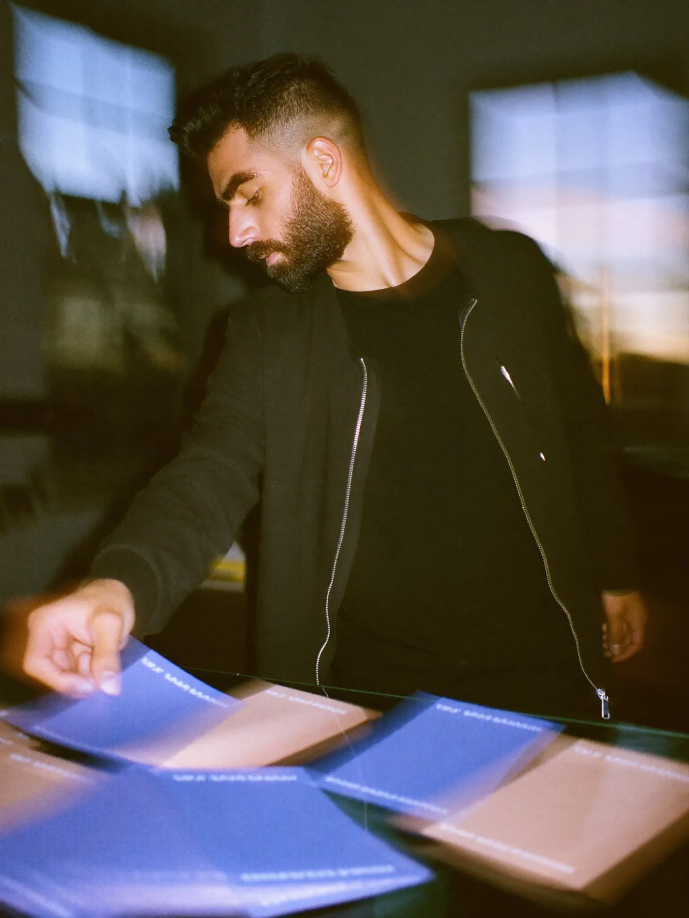 A bearded man in a charcoal jacket marking a stack of blush card beside a periwinkle stack in an office